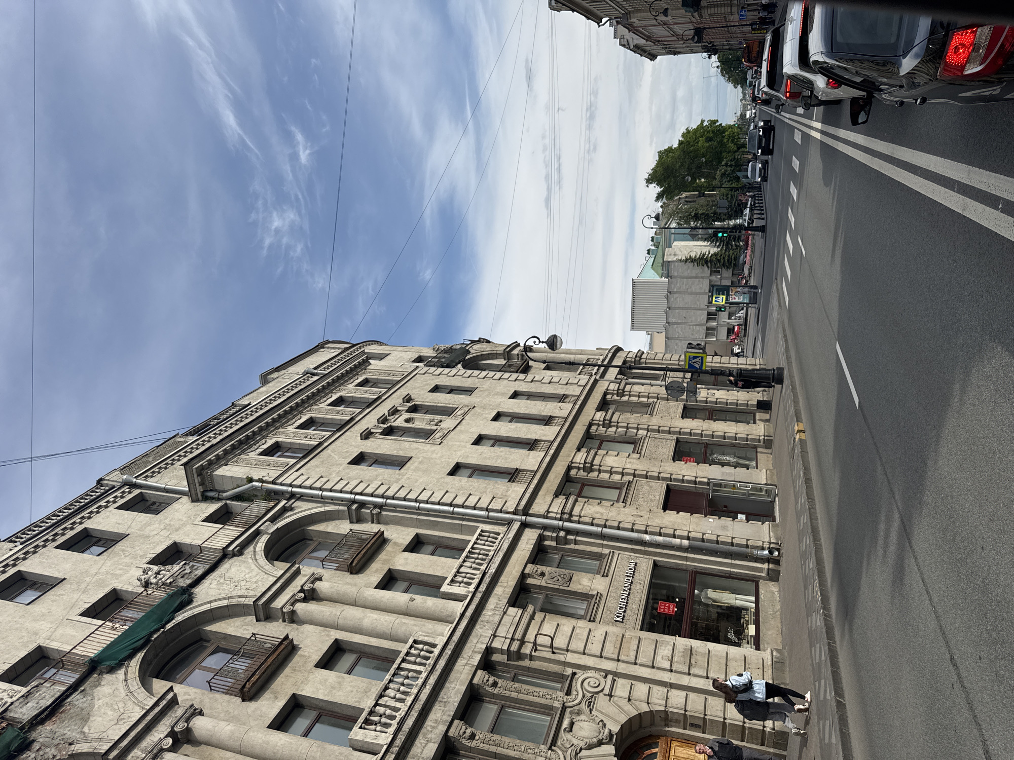 This media file captures an urban street scene in Saint Petersburg, Russia, under a bright blue sky with scattered wispy clouds, indicating daytime. The main subject is a large, ornate multi-story building on the left side of the frame. Constructed from light-colored stone or concrete, the building showcases a classical architectural style with decorative cornices, pilasters, and intricate window detailing. Some windows on the lower levels are arched, featuring decorative metal balconies or balustrades, while upper windows are rectangular. Dark-colored drainpipes or conduits run vertically down the facade.
Shopfronts are visible on the ground floor of the building. One prominent vertical sign reads "KUCHENLAND HOME," and another shop window displays a small red and white sign with "KGC LAND."
To the right, a wide asphalt road with clear double white lane markings stretches into the distance. Several cars, including a silver sedan and a red SUV, are visible, appearing to be in traffic. A pedestrian crosswalk is marked further down the street, and a green traffic light is discernible. A few pedestrians are seen walking on the sidewalk adjacent to the building, including one person in a light blue jacket. Overhead, multiple power or tram wires crisscross the sky. The scene depicts a typical busy city street on a clear day.