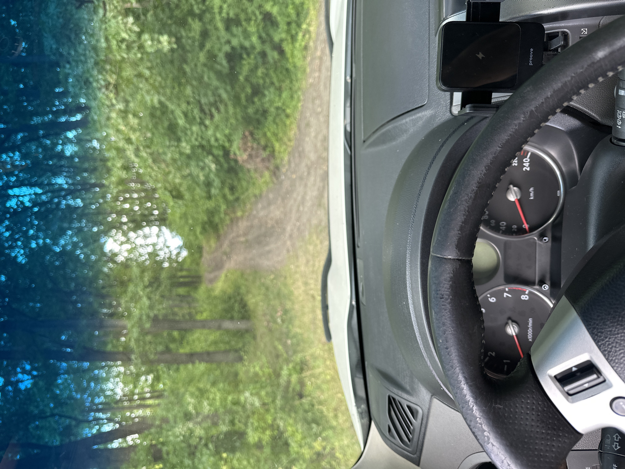The image shows the interior of a car, looking out through the windshield. The car appears to be on an unpaved road in a wooded area. The dashboard and steering wheel are visible in the foreground. Through the windshield, a dirt road winds through a dense forest with tall trees and green foliage. The sky above the trees is a bright blue. The scene suggests a journey through a rural or natural landscape, possibly in the city of Makiv, Ukraine, based on the provided context.