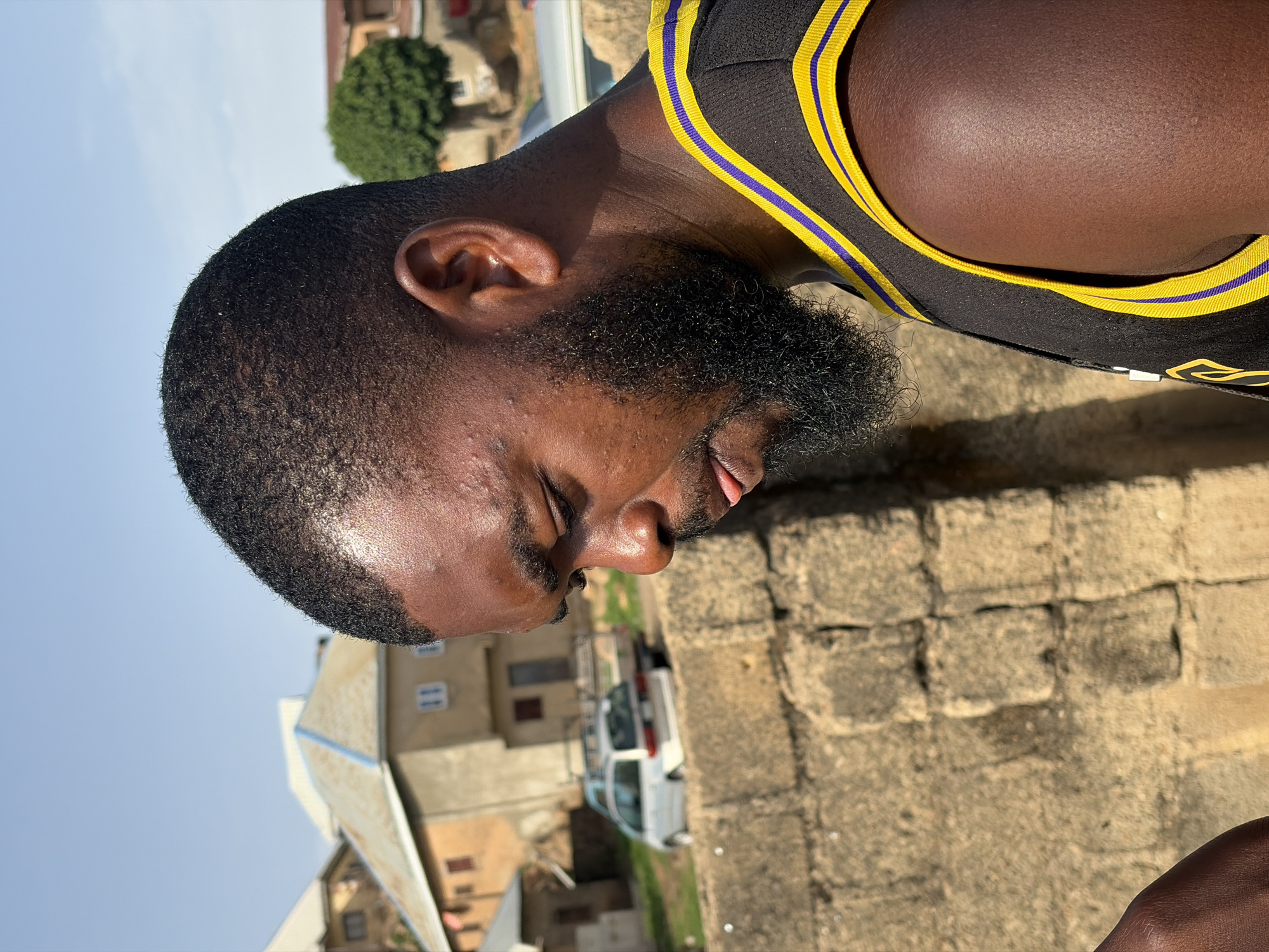 The image is an eye-level portrait of a dark-skinned man with a short, black, tightly-curled haircut and a full, dark beard. He is wearing a black tank top with yellow and blue trim along the neckline and armholes. His eyes are closed, and his tongue is slightly protruding from his mouth. The background includes a low, weathered brick wall and buildings in the distance, partially obscured by the angle of the shot. A white vehicle is also visible. The presence of the brick walls and surrounding buildings suggests an urban environment, possibly within the city of Jos, Nigeria.