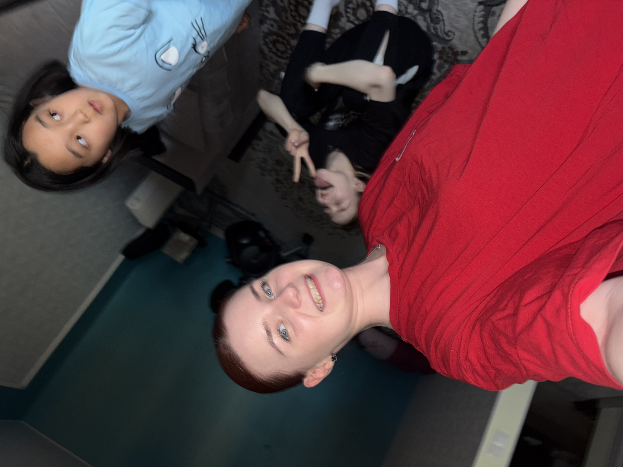 In this image, three individuals are visible in an indoor setting. Two are younger females, and one appears to be a young adult female. The setting includes a rug, a sofa, and walls. The lighting suggests it is nighttime or indoors with artificial lighting.
One of the younger females is lying on the rug, wearing a light blue long-sleeved shirt with a cat design. She is looking upwards and to the left. Beside her, another younger female is also on the rug, wearing a black shirt and black pants. She is making a peace sign with her fingers and sticking out her tongue, with her face turned towards the camera.
The third individual, a young adult female, is positioned prominently in the foreground, wearing a red t-shirt. She is smiling broadly at the camera, with her head tilted to the side. Her hair is reddish-brown, and she is wearing an earring.
The overall mood appears to be casual and playful. No visible text is present in the image. The context suggests a relaxed gathering or playtime among friends or family members.