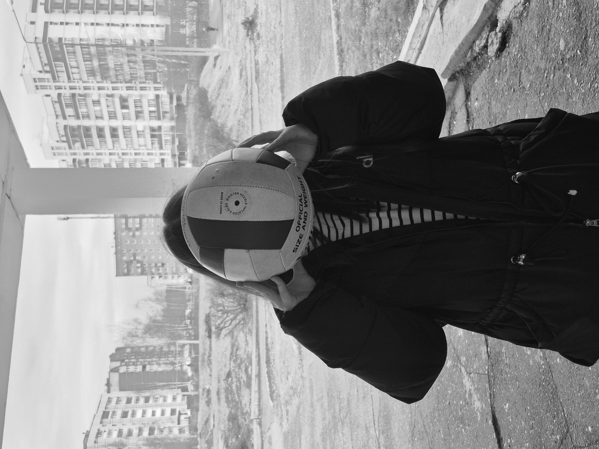 A person wearing a dark jacket and a striped shirt is holding a volleyball in front of their face. The volleyball is red and white and has the words "OFFICIAL SIZE AND WEIGHT" printed on it. The person is standing near a window, and through the window, buildings in the city of Chaikovsky, Russia, can be seen. The photo is in black and white. The ground appears to be asphalt.
