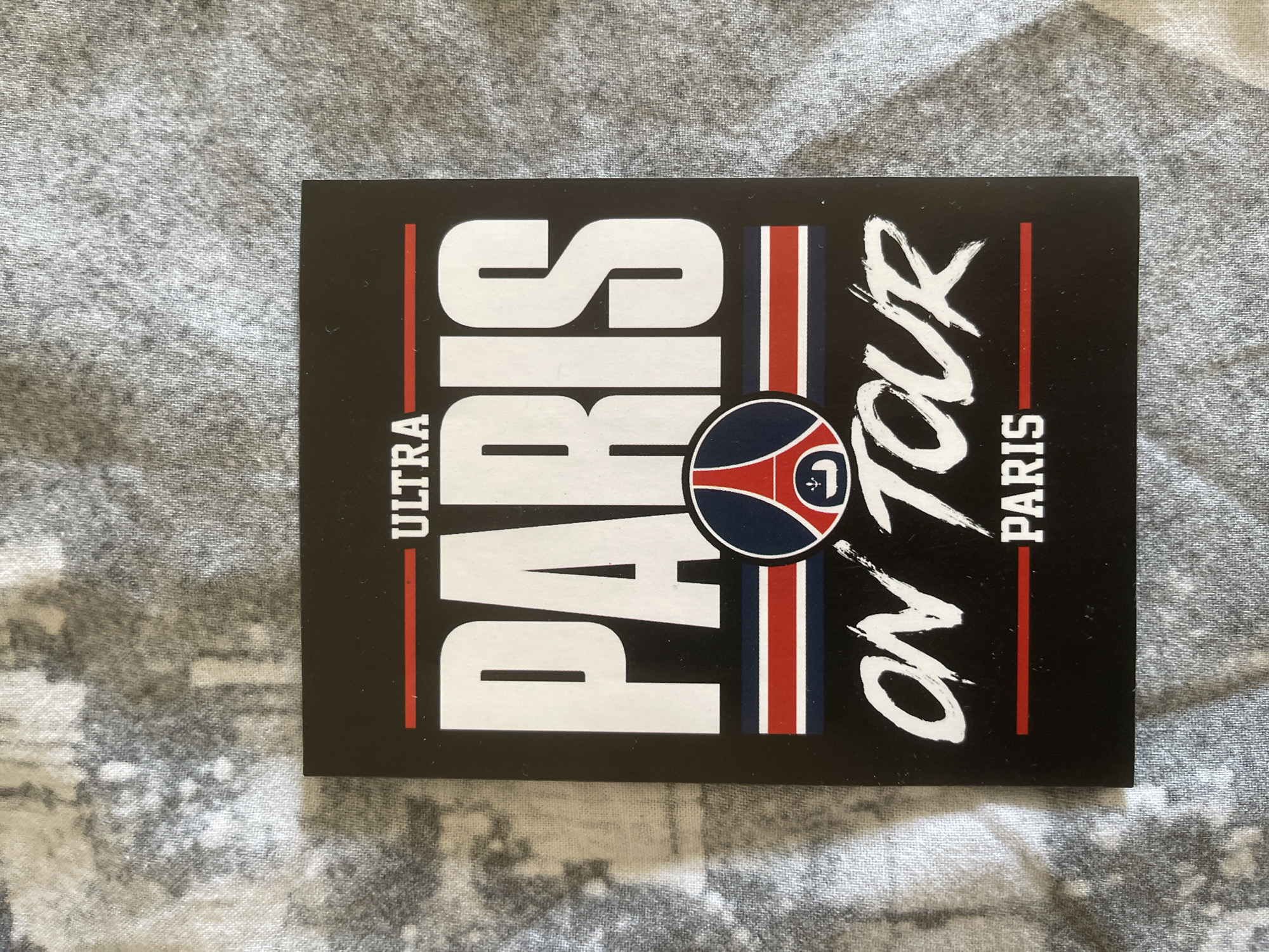 The image shows a square black sticker with white text and a crest. The text reads "ULTRA PARIS ON TOUR PARIS". The crest is circular and features a silhouette of the Eiffel Tower. The sticker is lying on a textured gray surface. There is no indication of the location in Paris, France.