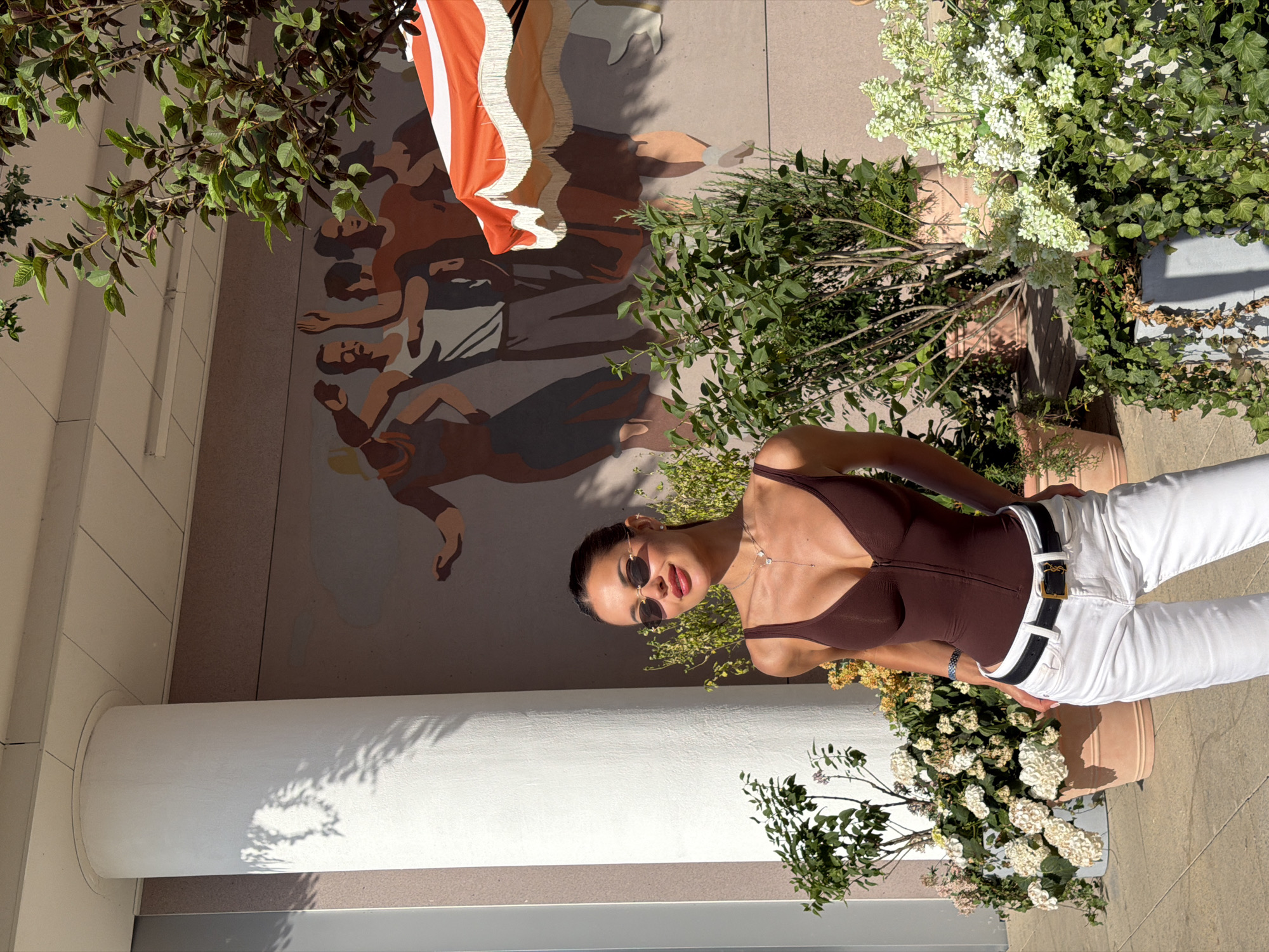 This vertical full-length shot captures a woman posing outdoors on a bright, sunny day, with a mural, lush greenery, and architectural elements forming the backdrop.
The main subject is a young woman, likely in her 20s or 30s, positioned slightly right of center. She has dark hair, wears dark, round sunglasses, and looks directly at the camera with a relaxed, slight smile. She is dressed in a form-fitting dark brown V-neck top or bodysuit, paired with white high-waisted jeans and a black belt featuring a prominent gold-toned "YSL" logo buckle. A delicate silver-colored necklace and a bracelet on her left wrist complete her accessories.
Behind her, a large mural on a building wall depicts stylized human figures in warm, earthy tones, appearing in various dynamic poses. Part of an orange and white striped fabric with fringe, possibly a decorative parasol or banner, is visible hanging over the upper portion of the mural.
The scene is surrounded by abundant green foliage, including tree branches overhead, shrubs with small white or light green flowers, and plants in terracotta pots. A large, white cylindrical column stands prominently on the left side, exhibiting distinct shadows from the overhead leaves, indicating bright sunlight. The ground appears to be paved, suggesting an urban outdoor environment such as a courtyard or plaza.
The bright sunlight and clear shadows confirm it is daytime, likely mid-morning or afternoon, with clear weather. The woman is posing for a photograph. The provided context places this scene in Moscow, Russia. The only visible text is the "YSL" logo on her belt buckle.