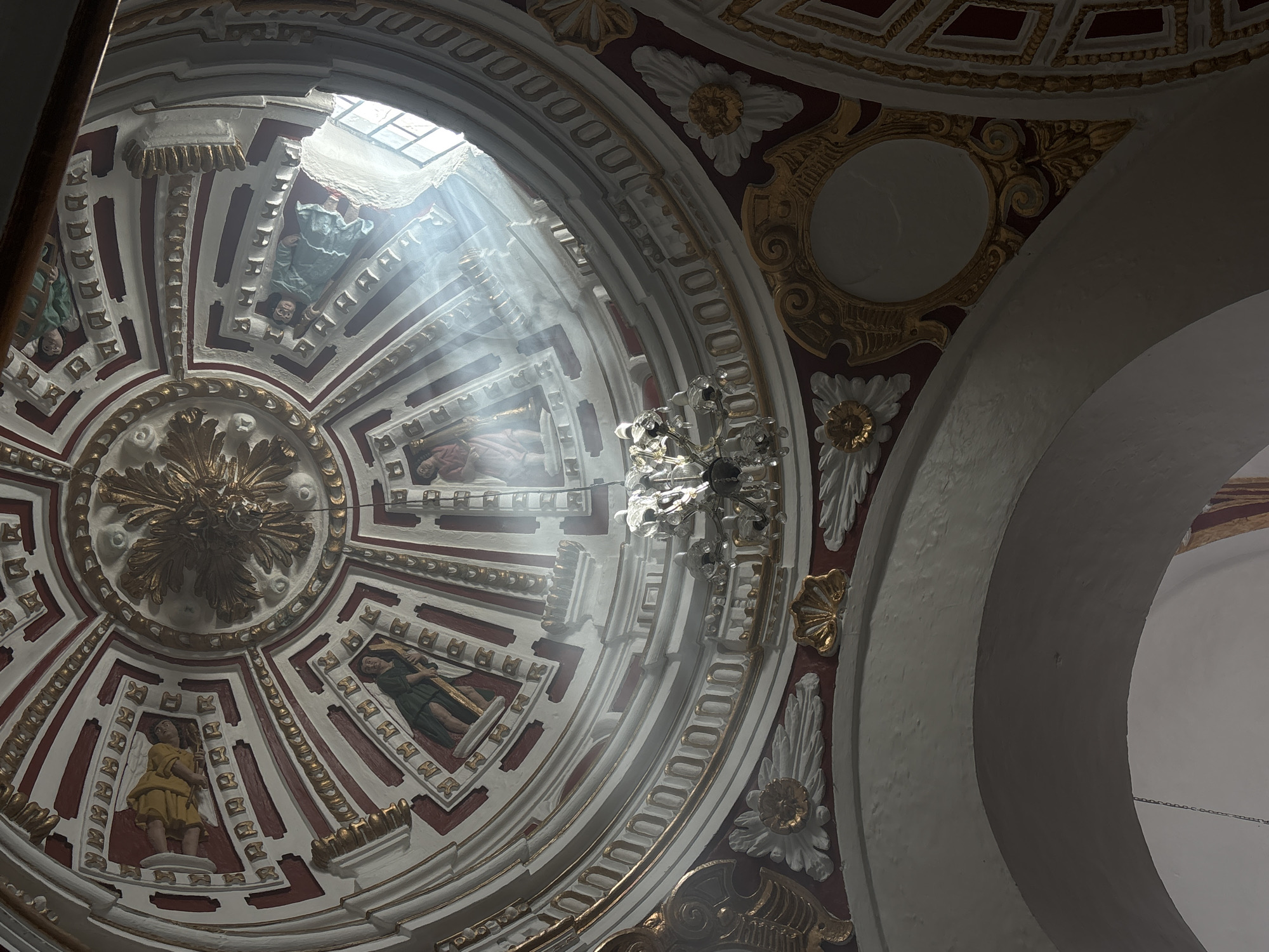 The image captures a view of the ceiling of a church in Puebla, Mexico. The ceiling is ornately decorated with a circular design. There are frescoes of religious figures painted within the geometric pattern. A central gold design is present at the center of the circular structure. Sunlight streams through an opening at the top, illuminating a chandelier hanging from the ceiling. The architecture is white and gold, with hints of maroon coloring the intricate designs.