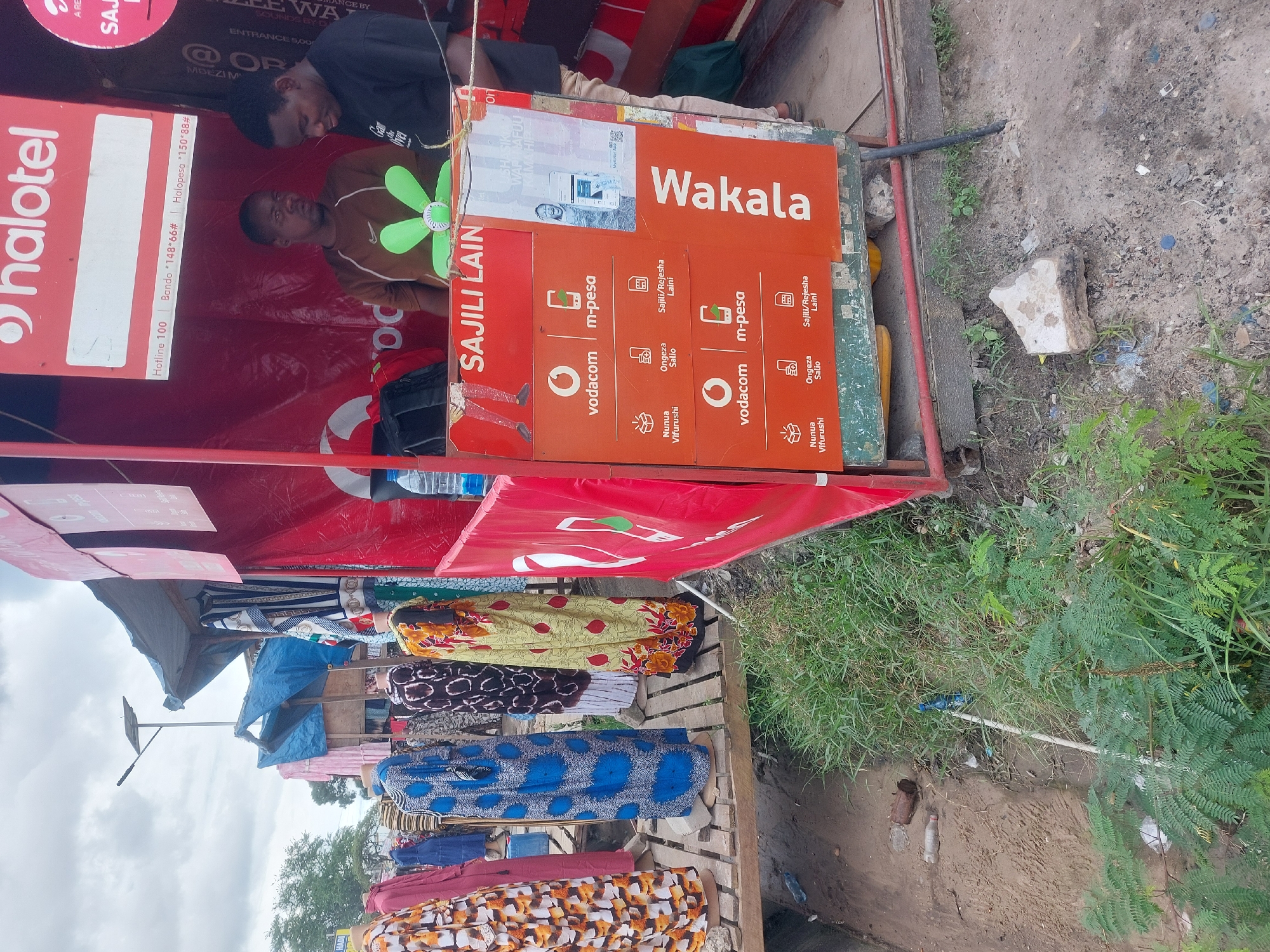 Here's a detailed description of the image:
**Scene:** A street-side business in what is likely Dar es-Salaam, Tanzania, based on the language and the presence of "Vodacom" and "Halotel" signage, both local telecommunications providers.
**Subjects:**
* **People:** Two men are visible near the front of the stall. One is wearing a brown shirt and is looking at the camera, and another in a black shirt.
* **Objects:**
* The stall is a makeshift structure with a red "Halotel" branded awning.
* "Vodacom" orange signage indicates a "Wakakala" (agent) for "M-pesa" (mobile money service). The signage mentions functions like "Sajili Lain" (Register Line), "Nunua Vifurushi" (Buy Bundles), and "Ongeza Salio" (Add Balance).
* Fabric/clothing is stacked for sale under the stall.
* Miscellaneous objects are visible on the ground near the stall.
**Environment:**
* The environment appears to be a semi-urban street with a dirt or unpaved roadside.
* Vegetation is present to the side of the stall.
**In summary:** This is a snapshot of a small business setup in Dar es-Salaam, Tanzania, providing telecommunication and mobile money services, with clothing for sale on the side. Two men are visible in the scene, presumably working at the stall.