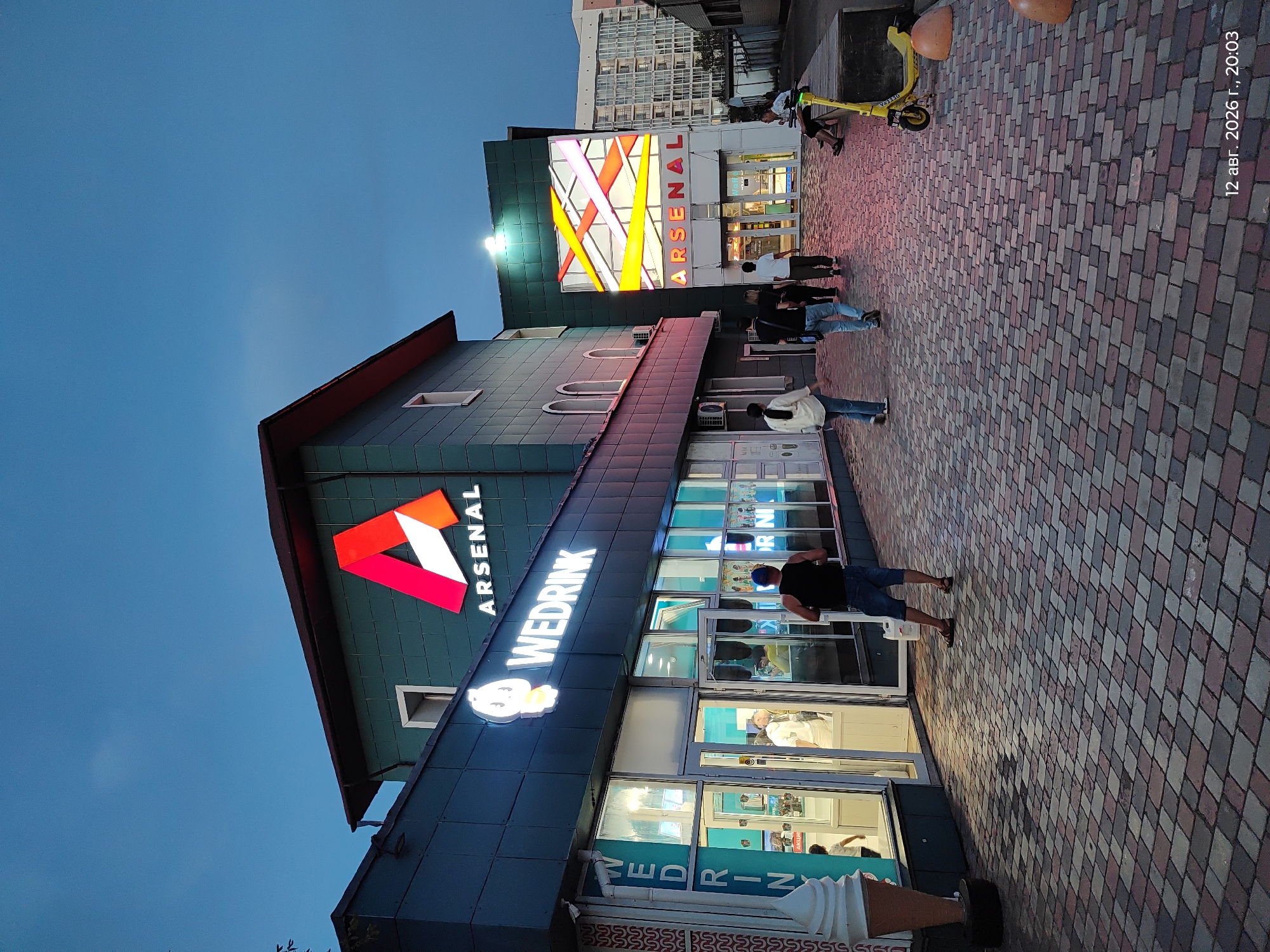 The image depicts an exterior view of a building with multiple businesses at dusk in Astana, Kazakhstan. The building features signs for "ARSENAL" and "WEDRINK." In front of the "WEDRINK" establishment, a large ice cream cone statue is visible. Several individuals are walking along a paved pathway in front of the businesses, some entering or exiting. A yellow electric scooter and two large orange bollards are situated to the right of the pathway. The sky is dark blue, indicating evening. A timestamp in the lower right corner reads "12 авг. 2026 г., 20:03," which translates to August 12, 2026, 8:03 PM.