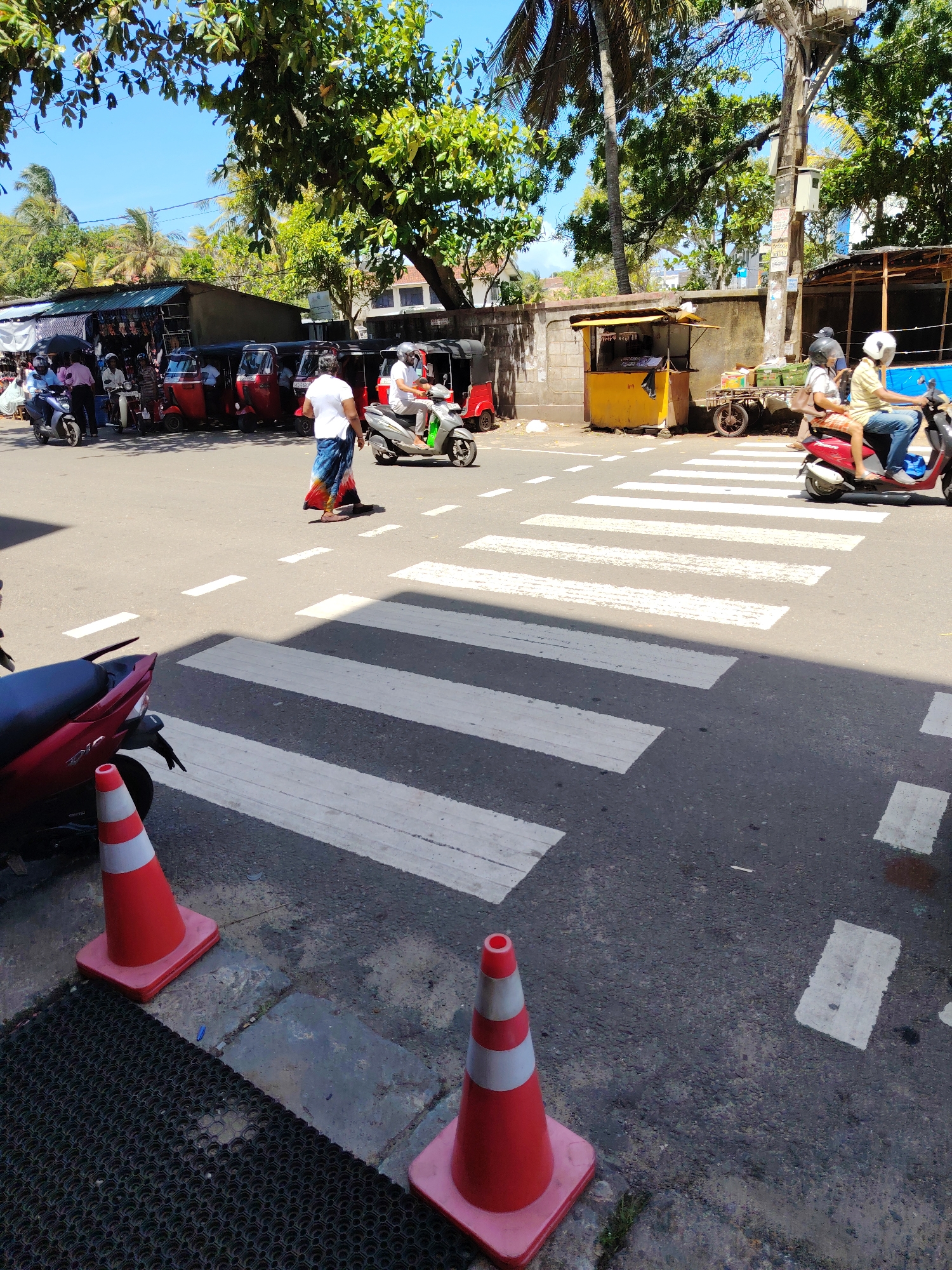 In Galle, Sri Lanka, on a sunny day, a busy intersection features a pedestrian crossing with white stripes. A red scooter is parked on the left, with an orange traffic cone nearby. Further into the road, a person in a white shirt and a colorful lungi walks across the pedestrian crossing. To the right, a person on a gray scooter, wearing a helmet and a light-colored shirt, crosses the street. Behind them, another person on a red scooter, also wearing a helmet, rides past. A small yellow kiosk with a yellow awning stands to the right of the road, with a cart carrying goods beside it. In the background, several tuk-tuks are parked, and shops with various goods are visible. Lush green trees and palm trees line the street, with a clear blue sky overhead.