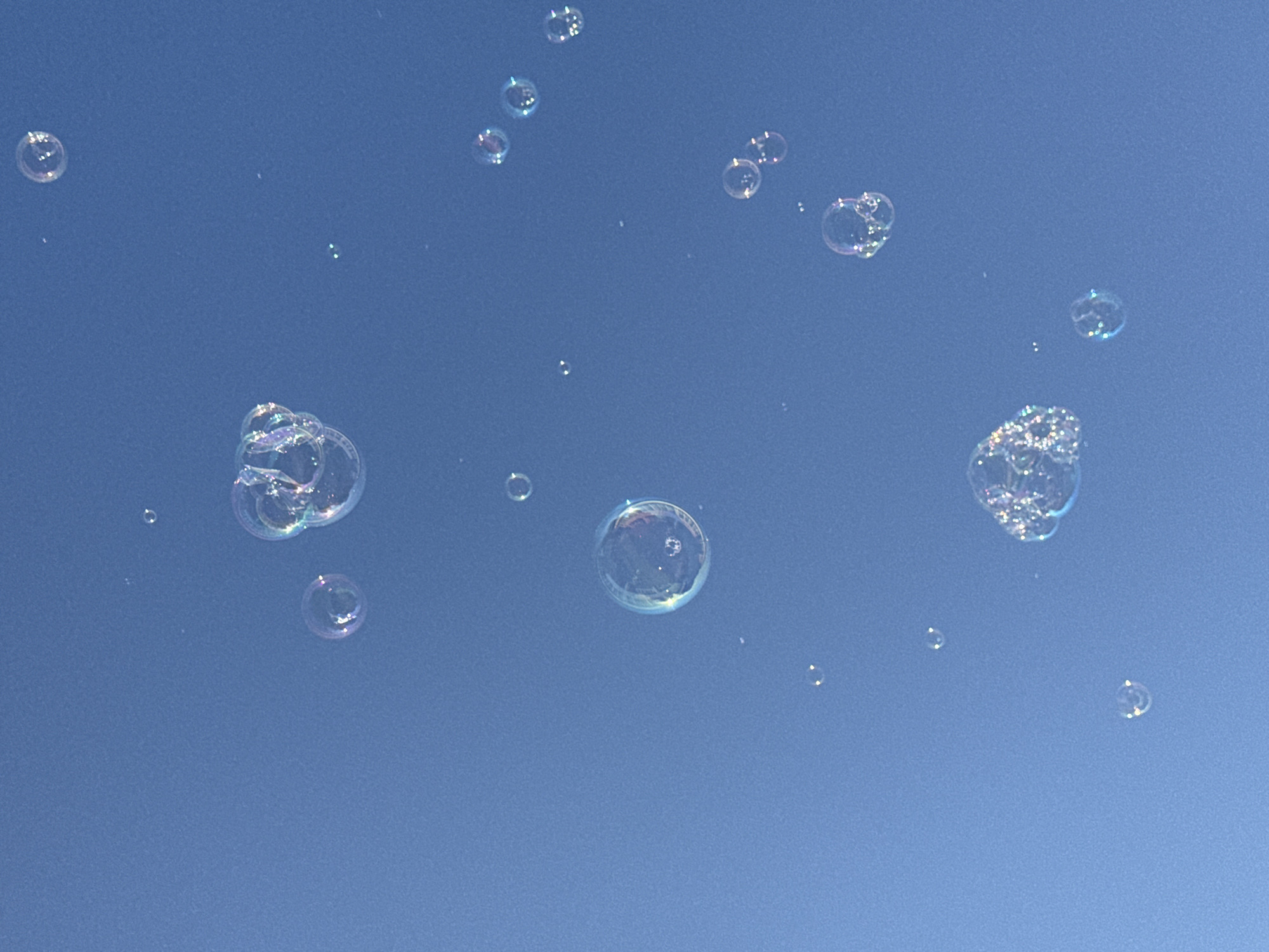 A collection of soap bubbles floats against a clear blue sky. The bubbles are of varying sizes and reflect light, displaying iridescent colors. The scene appears to be outdoors, with no specific Vityazevo, Russia landmarks visible. The focus is entirely on the bubbles and the sky.