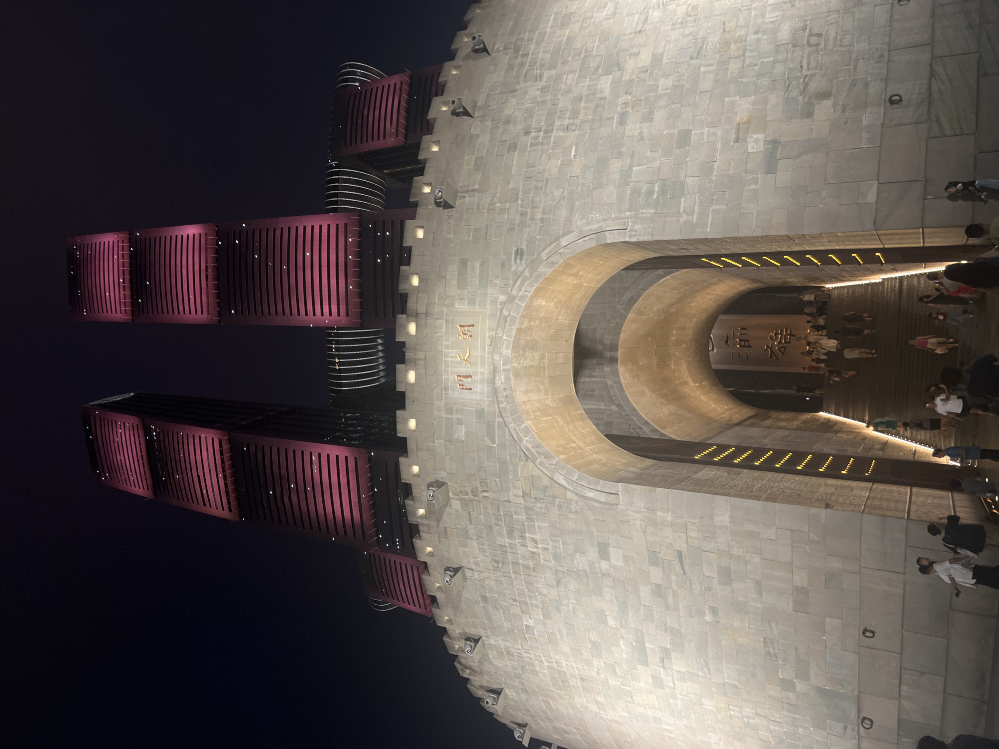 The image shows the exterior of a large, stone gateway structure at night, with two modern, illuminated skyscrapers in the background. The gateway has a massive arched opening leading to a staircase. The stone walls of the gateway are illuminated, and there are decorative elements and what appear to be gargoyles or decorative turrets along the top. The skyscrapers are tall and slender, with a distinctive design featuring horizontal bands of pinkish-red light. Chinese characters are visible on the facade of the gateway, reading "天 門 阙" (Tianmen Que). People are seen walking up the stairs within the gateway and gathered in front of it. The overall environment is dark, indicating nighttime, and the lighting emphasizes the architecture of the gateway and the illuminated skyscrapers. This location is identified as the Tianmen Que in Nan'an District, China.