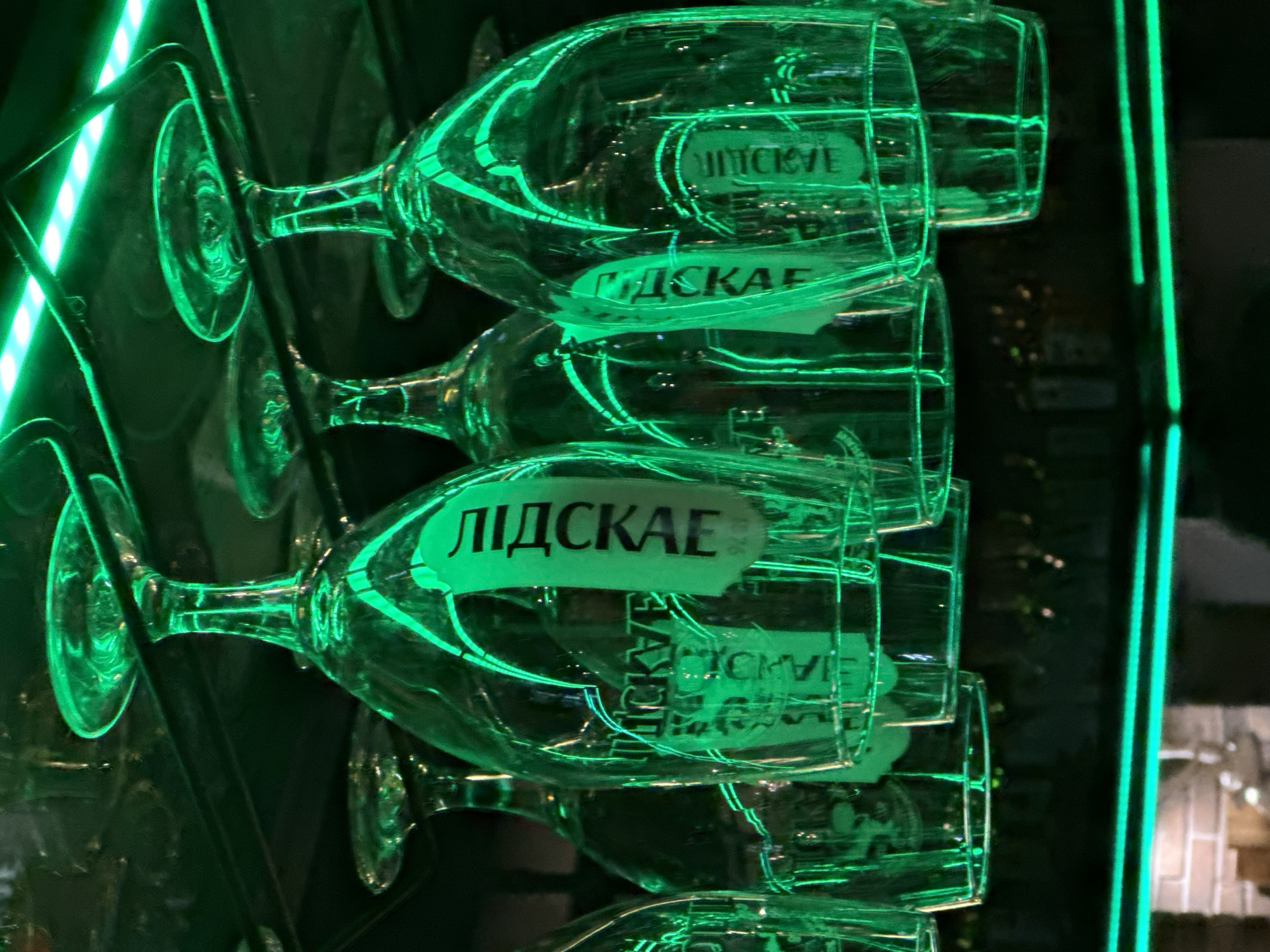 In Minsk, Belarus, this image captures a close-up view of several clear wine glasses hanging upside down from a dark metal rack. The glasses are illuminated by a vibrant green neon light, which creates strong reflections on their surfaces. Two of the glasses have visible paper labels with black Cyrillic text. The text on one label appears to read "МІДСКАЕ" and on another "МІДСКАЕ," suggesting they are from a brand or establishment. The background is dark and out of focus, with hints of shelves and possibly bottles, implying the setting is a bar or restaurant. The overall scene is dimly lit, with the neon green light being the dominant source of illumination, casting an atmospheric and somewhat futuristic glow. There are no people visible in the image.