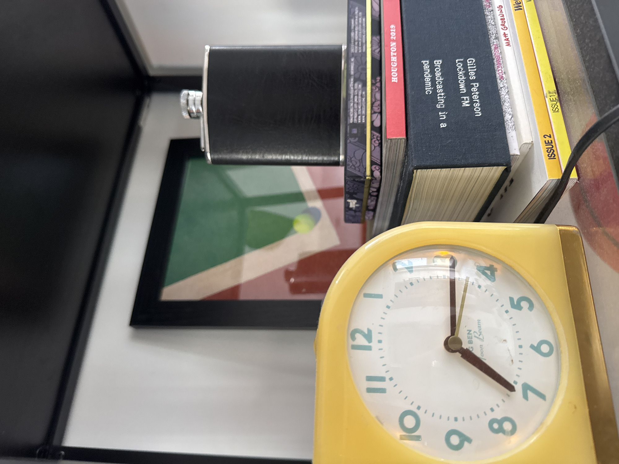 The image presents a still life arrangement. A yellow alarm clock, showing the time around 4:35, is in the foreground. Stacked on top of the clock are several books, including one with the title "Broadcasting in a pandemic" and another titled "Houghton 2019." A silver flask is positioned on top of the books. In the background, a framed image containing green and red geometric shapes is partially visible. The setting appears to be indoors, possibly on a shelf or desk.