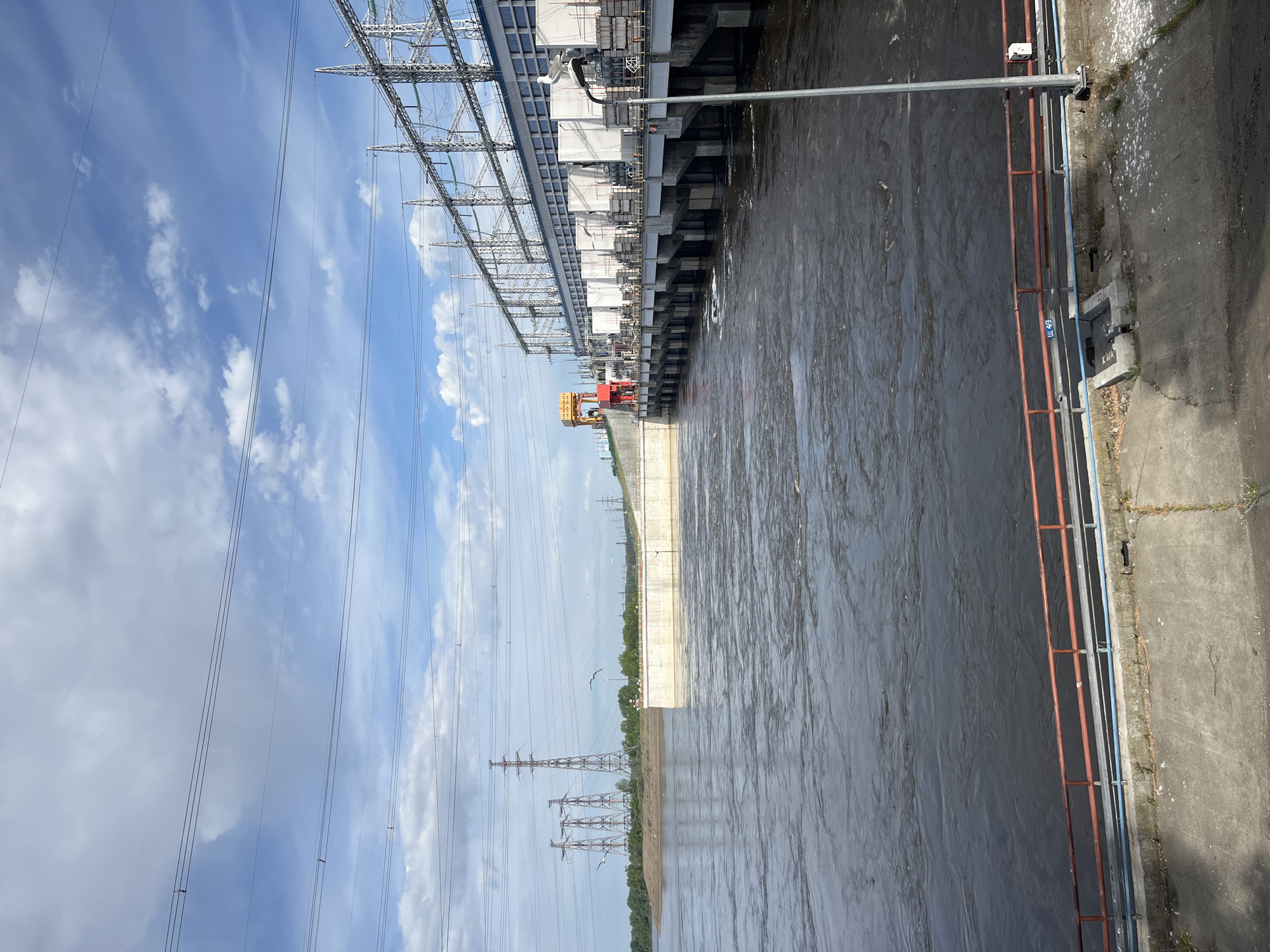 This image shows the Votkinsk Hydroelectric Station in Chaikovsky, Russia. It depicts a wide river with turbid water flowing along a concrete embankment. On the left, a power substation with metal framework, electrical transformers, and power lines is visible. A large red gantry crane or lifting mechanism is located near the water's edge. Transmission towers extend into the distance. The sky is partly cloudy. A metal railing runs along the edge of a concrete platform on the right side of the frame.