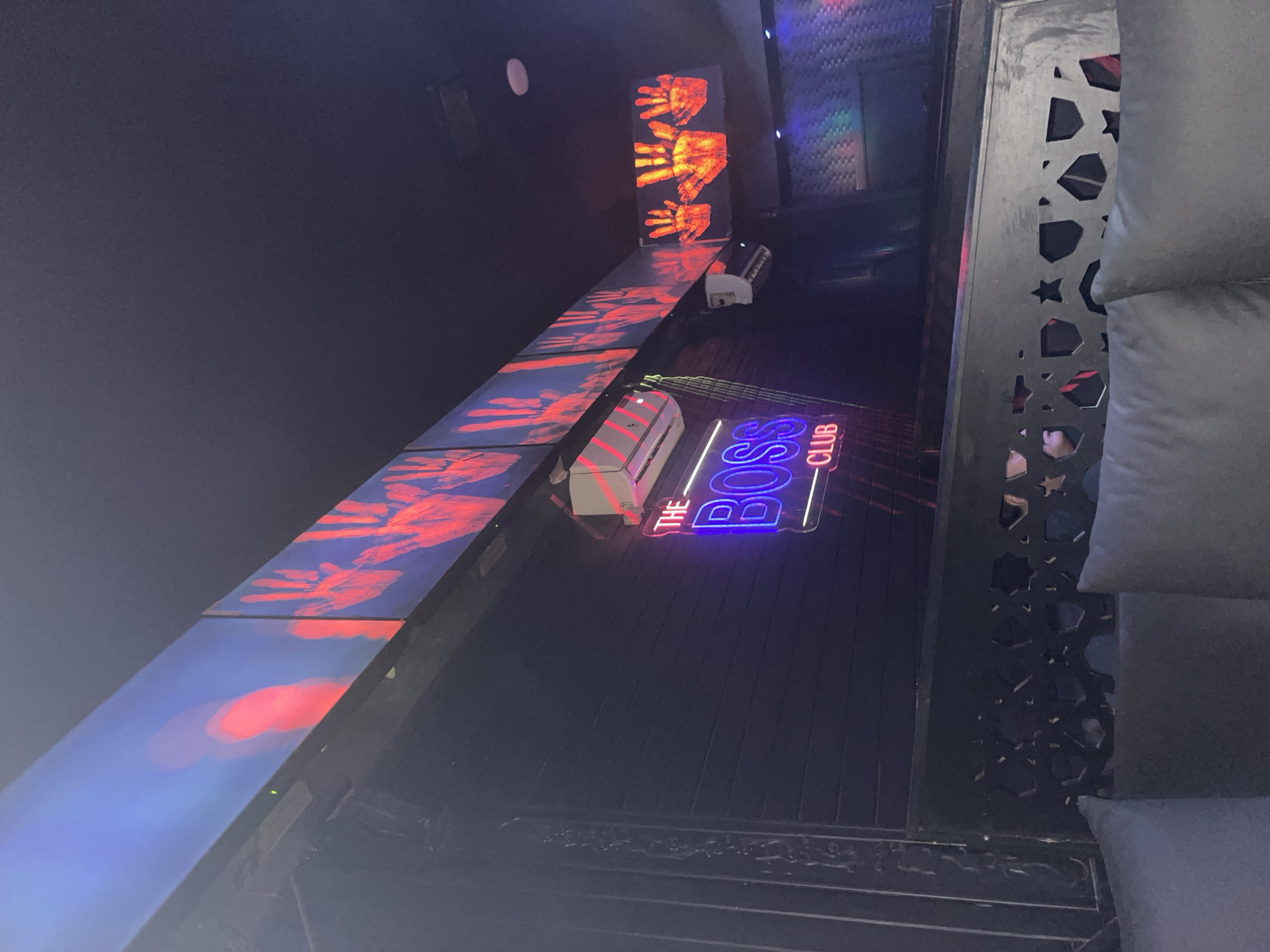 The image shows the interior of a dimly lit establishment with neon signage and decorative elements. In the center, a neon sign displays "THE BOSS CLUB" in a vibrant blue and pink glow. Above the sign, a row of screens or panels shows animated red handprints against a dark blue background. To the right, a similar display of red handprints is visible. There are air conditioning units mounted on the wall. In the foreground, dark gray upholstered seating is visible, separated by a decorative metal partition with a geometric pattern. The overall ambiance suggests a nightclub or entertainment venue. The location is identified as Amman, Jordan.