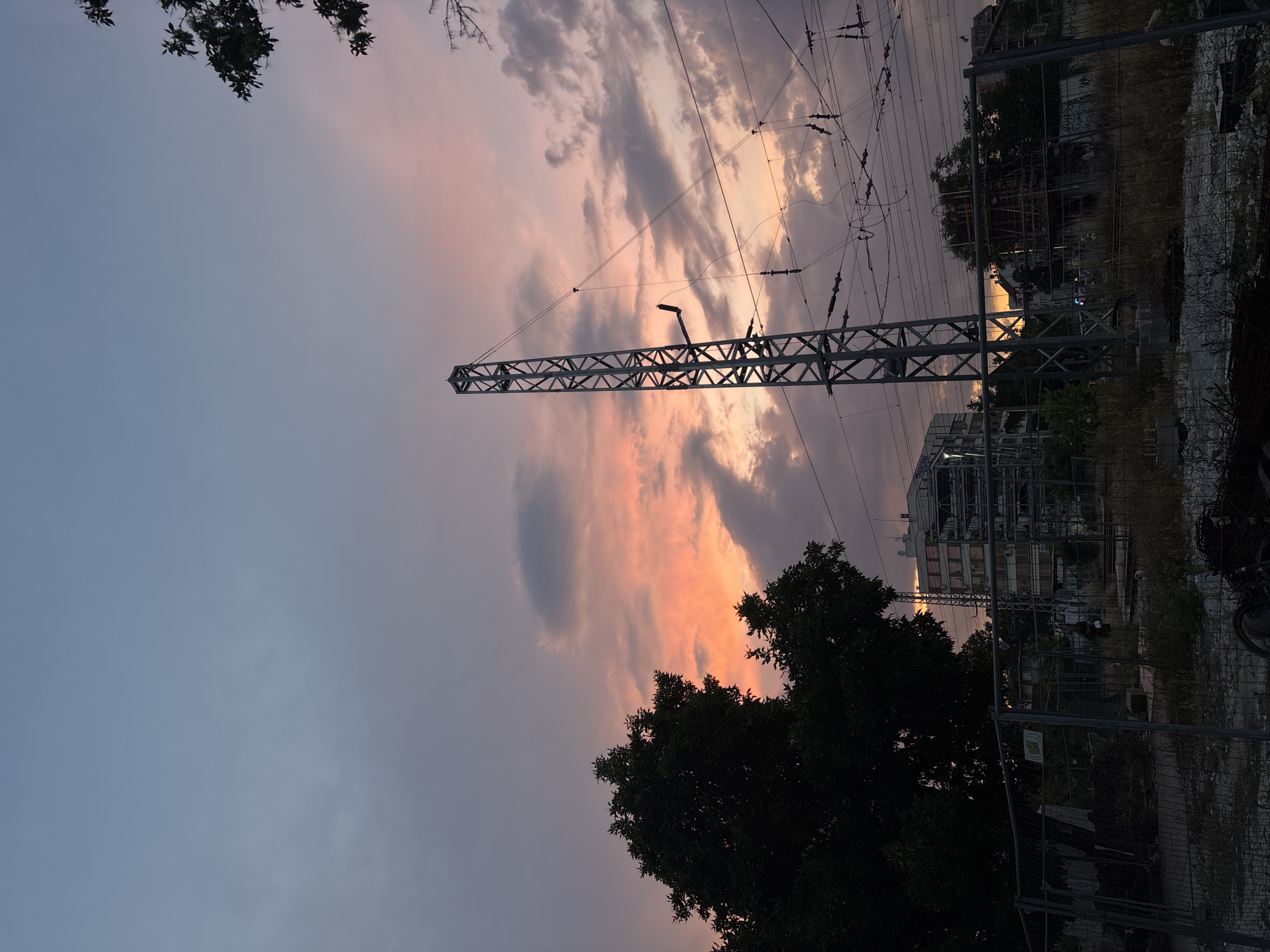 The image captures a dusk scene with a dramatic sky featuring hues of pink, orange, and grey clouds. In the foreground, a chain-link fence encloses a construction site or an undeveloped area with debris and dry vegetation. Dominating the mid-ground is a tall, lattice-like metal pylon, likely an electricity transmission tower, with numerous wires suspended from it. Behind the pylon, a multi-story building with scaffolding is visible, suggesting ongoing construction or renovation. Silhouetted against the sky on the left is a large, dense tree. The overall impression is an urban landscape at the edge of development, with industrial infrastructure juxtaposed against natural elements and a vibrant sky. While specific landmarks in Plovdiv, Bulgaria, are not explicitly identifiable from the image alone, the scene is consistent with areas of industrial development or transportation hubs within the city. No people are discernible in the image.