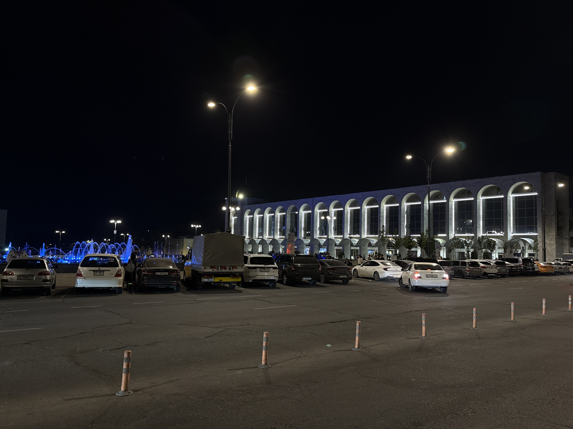 This is a nighttime, eye-level, outdoor, wide shot of a public square in Bishkek City, Kyrgyzstan. The scene captures a building with a long, arched facade illuminated by interior lights, creating a pattern of light and shadow. Several streetlights with multiple bulbs are visible, casting pools of light on the asphalt. In the foreground, there is a paved parking lot with multiple cars parked and driving. Orange and white bollards are spaced across the lot. To the left, a large fountain with blue illuminated water jets is active. The sky is dark and clear.