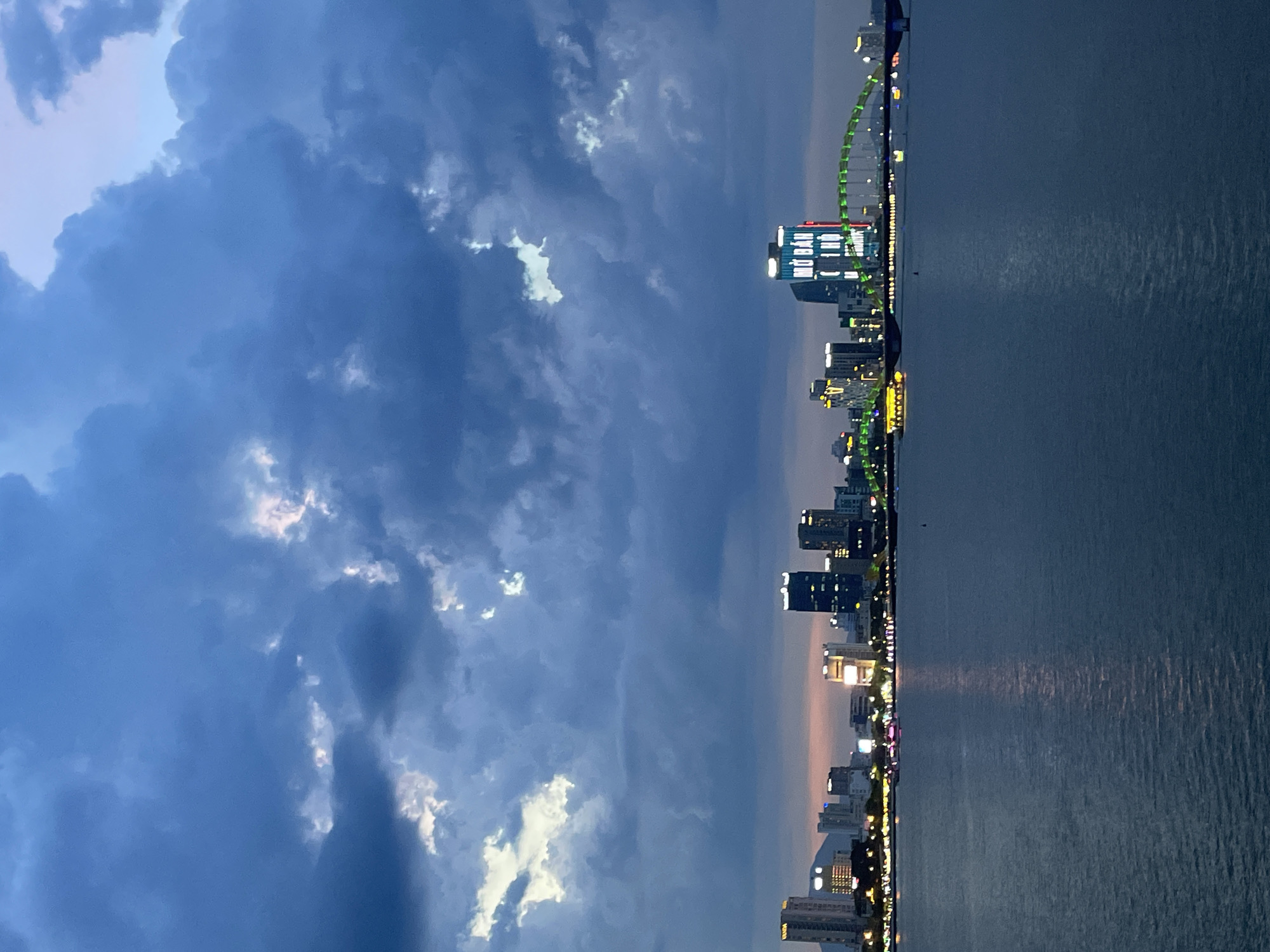 The image depicts a panoramic view of the Đà Nẵng cityscape at dusk. The scene is divided into two distinct halves: the sky on the left and the city and water on the right.
The left side is dominated by a dramatic sky filled with dark, heavy clouds that are beginning to break, revealing patches of lighter sky and some illuminated clouds. The interplay of light and shadow suggests a transition from day to night, possibly after a storm.
The right side showcases the Đà Nẵng skyline, characterized by a series of modern buildings illuminated with colorful lights. Prominently featured is a large, arched structure, likely a bridge, that glows with green lights. The city lights are reflected on the calm surface of the water, creating a shimmering effect. The shoreline is lined with more lights from buildings and possibly streetlights, giving the impression of a vibrant urban environment. A few small boats or buoys are visible in the water.
The time of day is evident as twilight, with the sky transitioning from daylight to darkness, and artificial lights of the city becoming prominent. The weather appears to be clearing after some cloud cover. The location is clearly Đà Nẵng, Vietnam, with the distinctive architecture and waterfront view being characteristic of the city. There is text visible on one of the buildings, appearing to be Vietnamese characters, though not entirely legible. The overall mood is one of urban beauty and transition, with the dramatic sky juxtaposed against the illuminated city.