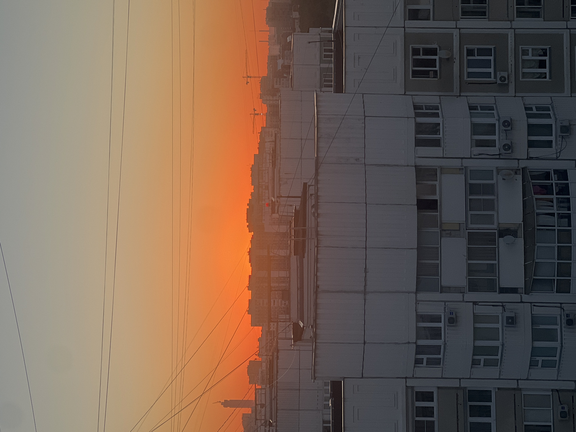 The image depicts a cityscape at sunset, likely in Moscow, Russia. The sky is dominated by a vibrant orange and yellow gradient, indicative of the sun setting or rising. Several multi-story residential buildings, characteristic of Soviet-era architecture, are visible in the foreground and mid-ground. These buildings are primarily concrete structures with numerous windows and balconies. Overhead, power lines crisscross the sky, a common sight in urban environments. In the background, a taller building with a distinct silhouette and construction cranes suggest ongoing development or a prominent landmark. The overall impression is one of a densely populated urban area bathed in the warm, dramatic light of the twilight sky. There are no visible people or animals.