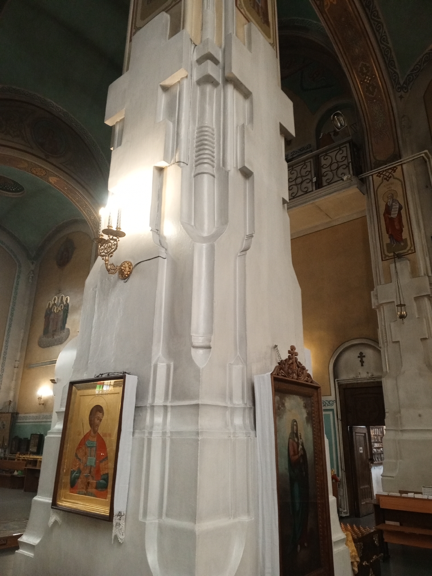 The photograph is an interior view of a religious building, likely a church or cathedral, with a focus on a large, ornate pillar in the foreground. The pillar is predominantly white with geometric carvings and vertical fluting. To the left of the pillar, a framed icon depicting a person in red and blue robes with a halo is visible, leaning against the pillar and draped with white fabric. To the right, another framed icon, taller and narrower, shows a woman in a blue and red garment holding a child. This icon is also draped with white fabric.
In the background, a balcony with decorative metal railings is visible on an upper level. The architecture suggests a classical or Byzantine influence, with arched ceilings and ornate decorative elements. Various religious icons and paintings are placed on the walls, and a chandelier hangs from the ceiling. The lighting appears to be artificial, casting warm glows on the surfaces. The overall impression is one of solemnity and reverence, typical of a religious space. The location is unknown.