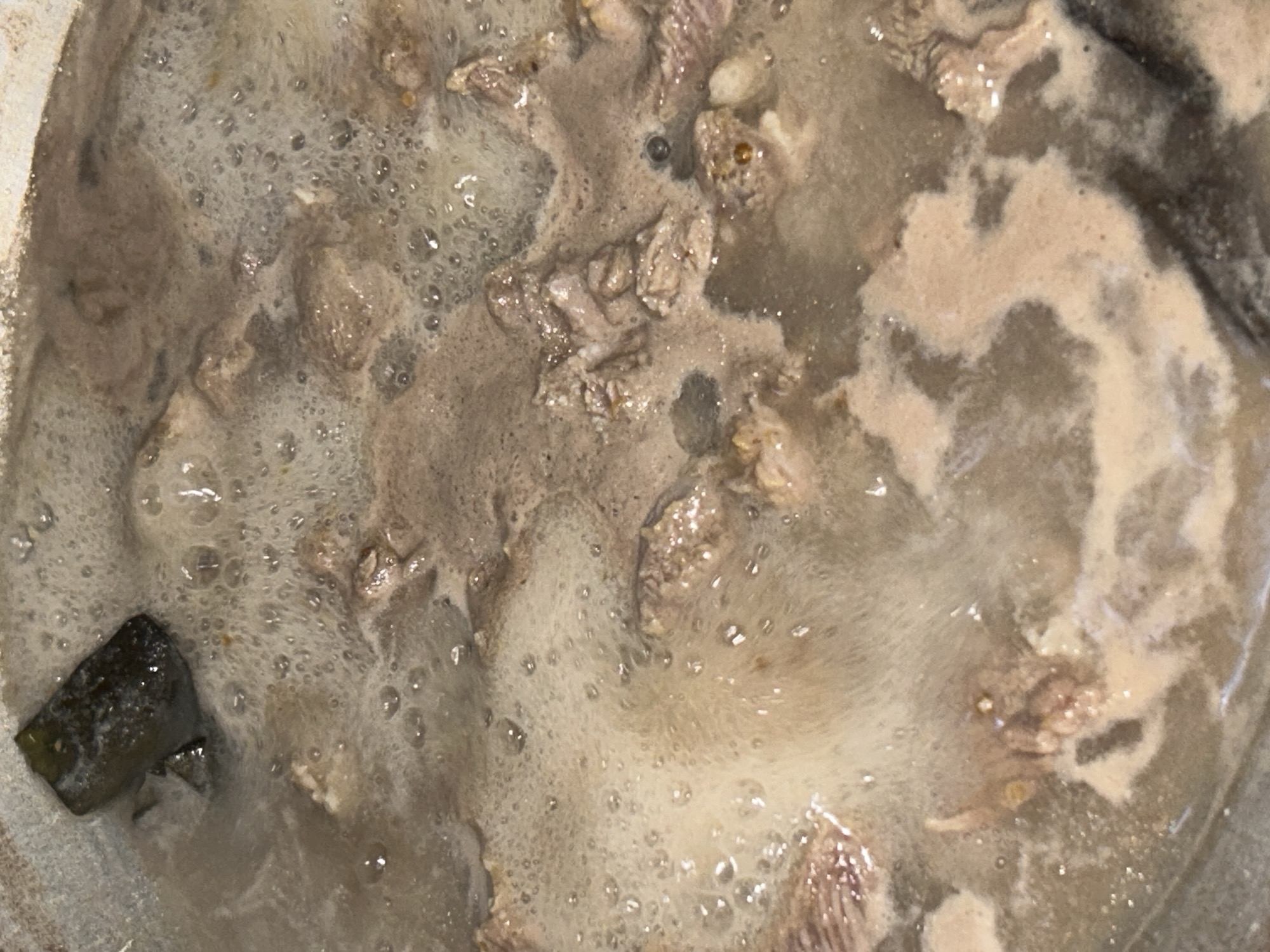 The image shows a close-up view of what appears to be a pot of stew or soup. The liquid is a cloudy, grayish-brown color with numerous bubbles on the surface. There are small pieces of meat or other ingredients visible throughout the mixture. A dark green chunk is visible in the bottom left corner of the image. There is no visible location data or information about the city of Lhokseumawe, Indonesia in the image.