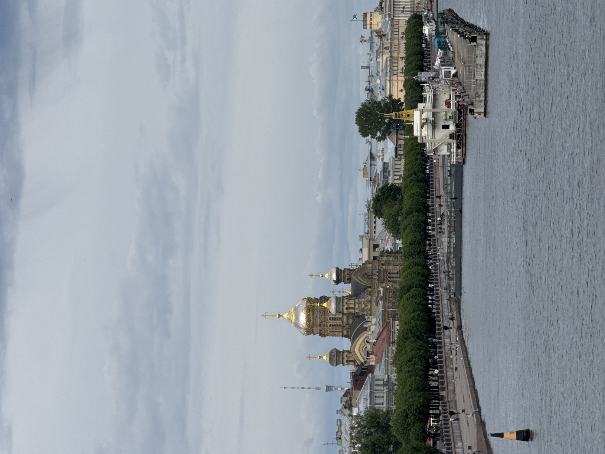 This is a medium-shot, eye-level, outdoor, wide-angle, natural light photograph of a cloudy day over the Neva River in Saint Petersburg, Russia. On the left side of the frame, a large, multi-domed Orthodox church with golden spires dominates the skyline. Behind the church, a tall, red and white striped radio tower is visible. In the middle ground, a tree-lined embankment runs along the river, with people walking and standing. A concrete pier with a white building and several docked boats extends into the river on the right side of the frame. A black and yellow buoy is visible in the foreground of the river. The water is dark and choppy, and the sky is overcast with gray clouds.