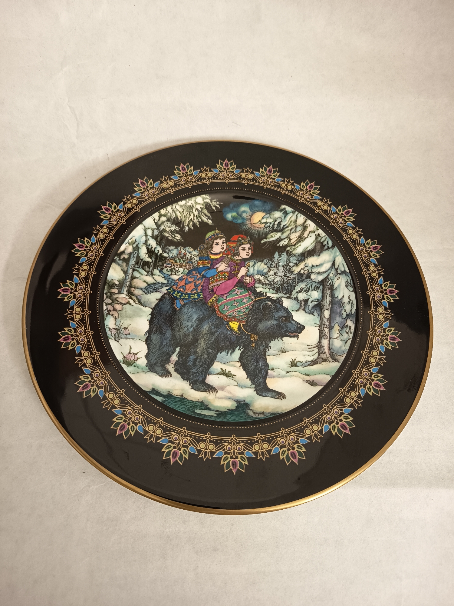 The image shows a decorative plate. The plate features a central illustration of two figures riding a black bear through a snowy forest. The figures are dressed in colorful, ornate clothing. The forest is depicted with snow-covered trees and a body of water running through it. In the background, there's a depiction of a castle or village under a night sky with a moon. The plate itself is black with a gold-colored rim, and a detailed border containing decorative floral patterns with blue and purple accents. The plate is lying on a white surface. There is no indication that the image was taken in the city of Wangen bei Olten, Switzerland.