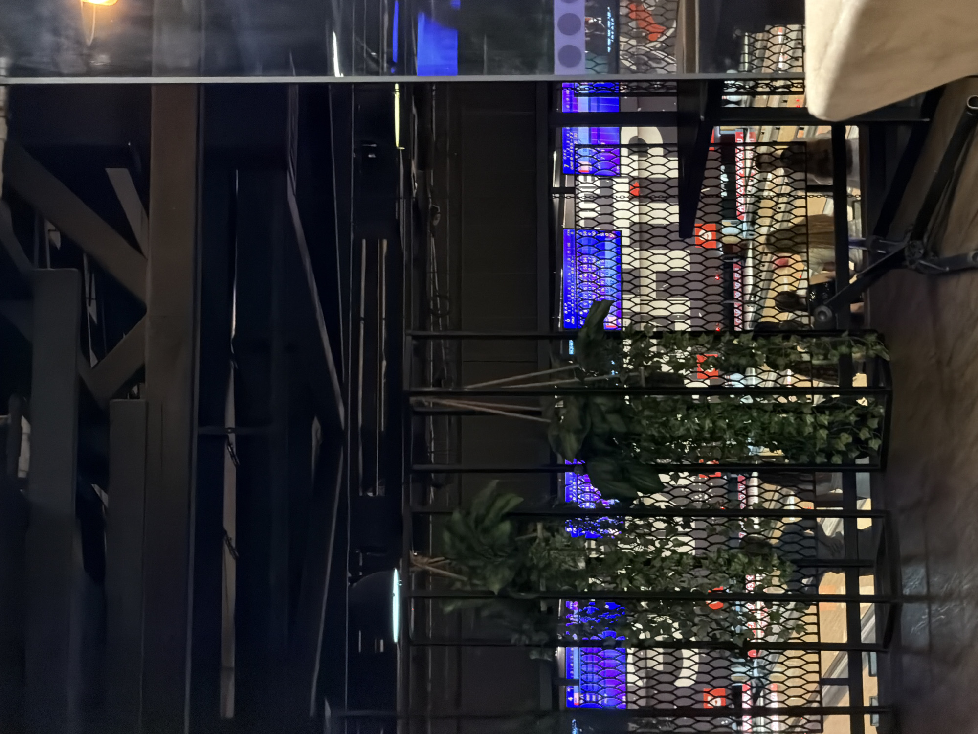 An indoor bowling alley is depicted. The image is framed by black metal structures and hanging artificial plants. In the background, bowling lanes are visible with glowing purple and blue screens displaying scores. Several people are seated at tables or walking near the lanes, their faces obscured by the distance and lighting. The environment appears modern and dimly lit, with spotlights and screens providing illumination. The location is identified as Kaliningrad, Russia.