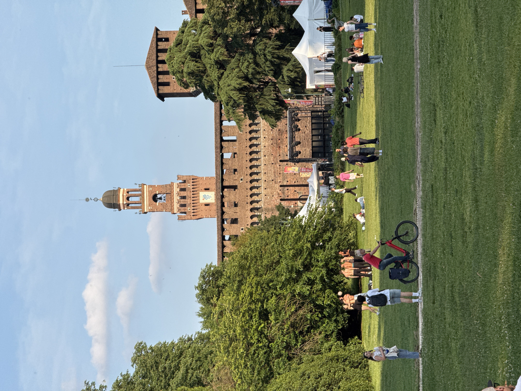 This image captures a wide view of Parco Sempione in Milan, Italy, with the Castello Sforzesco dominating the background. The time of day appears to be late afternoon or early evening, evidenced by the warm light and long shadows cast across the green lawn. The sky is light blue with sparse white cumulus clouds.
In the background, the red brick walls and towers of the Castello Sforzesco are clearly visible. The prominent Filarete Tower stands centrally, featuring a dark dome, a clock face, and arched openings. To its right, a shorter, square tower with a distinct roof is present. The castle walls exhibit battlements and arched openings. In front of the castle walls, an event area is set up, featuring multiple white canvas tents and a performance stage equipped with black speakers and lighting rigs, enclosed by a black metal fence.
The mid-ground and foreground comprise a vast expanse of green grass with a dirt path running horizontally across the center. Dense green trees occupy the left side of the frame, with additional trees scattered throughout the park.
Approximately 30-40 people are dispersed across the scene. On the dirt path, from left to right, a woman in light jeans and a white top stands holding a white bag, a man with a black backpack stands facing away, and a man in a red shirt rides a red bicycle equipped with a black pannier bag. To the left of the path, under a tree, a group of approximately five young adults, some shirtless, are engaged in playing with a white ball. Other individuals are seen walking on the grass, including a group of three men further left. In the mid-ground, near the event tents, several groups of people are standing or sitting on the grass. A woman with blonde hair, wearing a black top and light-colored jeans, stands facing left near the center-right. Further groups of people are scattered throughout, relaxing or moving through the park.