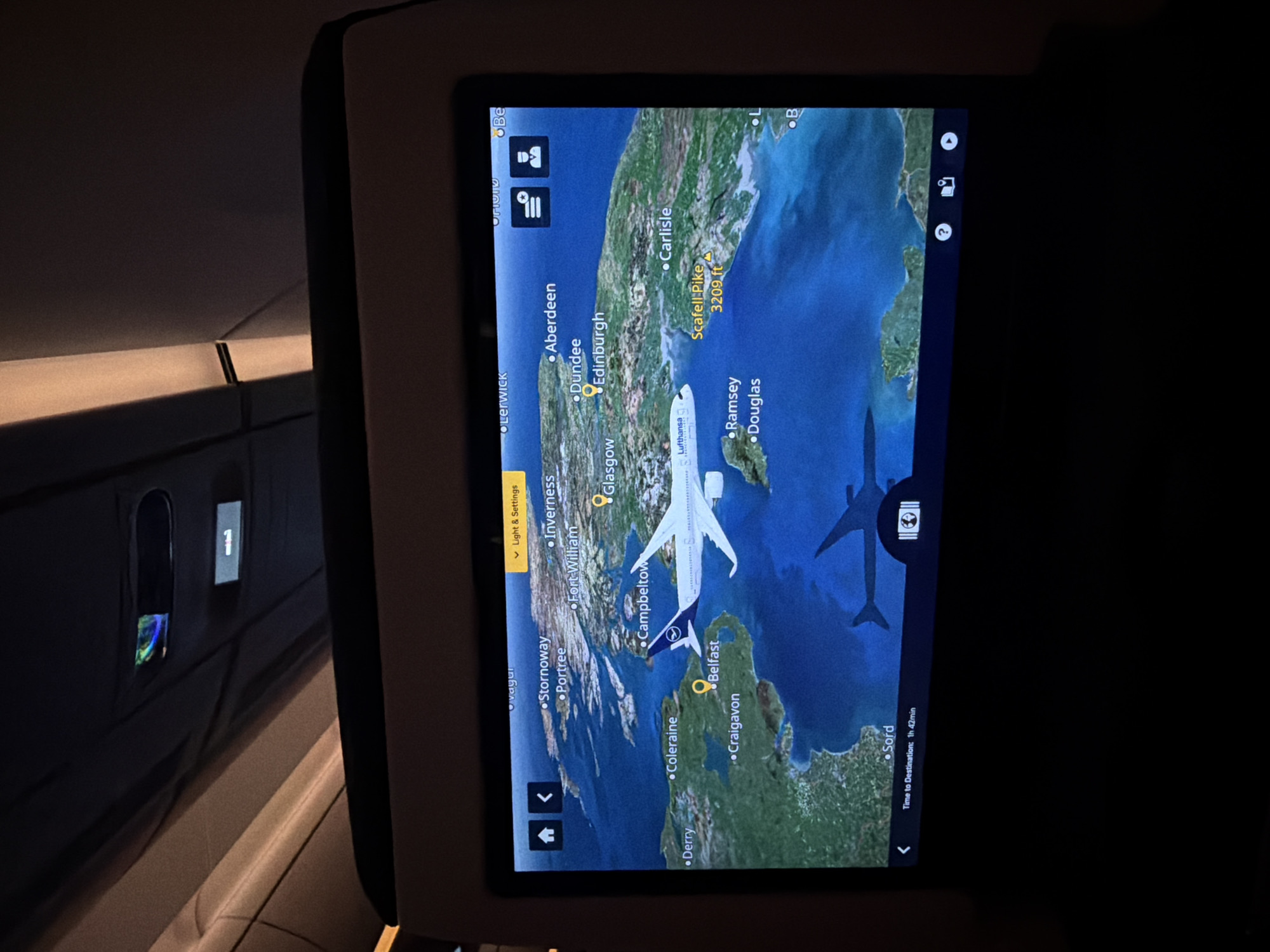 The image captures an in-flight entertainment screen displaying a real-time flight map within a darkened airplane cabin. The screen, held by a seat-back mount, shows a geographical view centered over the British Isles, specifically detailing parts of Scotland, Northern Ireland, the Isle of Man, and northern England.
On the map, a digital representation of a Lufthansa airplane is depicted, indicating its current position over the Irish Sea, east of Belfast and west of Carlisle. Key cities and landmarks labeled on the map include Stornoway, Portree, Inverness, Fort William, Campbeltown, Coleraine, Derry, Belfast, Craigavon, Glasgow, Edinburgh, Dundee, Aberdeen, Carlisle, Scafell Pike (3209 ft), Ramsey, and Douglas.
Visible text on the screen includes "Lufthansa" on the plane model, "Time to Destination: 1h 42min", and a destination or waypoint labeled "Sord". A button labeled "Light & Settings" is also visible in the top left. Other UI elements include various icons for navigation, settings, and information, along with partially visible text like "Lerwick," "vayur," "Be," "L," and "B."
The cabin environment is dim, suggesting nighttime or a dark cabin setting. To the left, a window shade is visible, with a sliver of light illuminating the area beneath it. Below the window, part of the cabin wall with a small illuminated panel or screen is discernible. The overall scene indicates a passenger's view during an active flight.