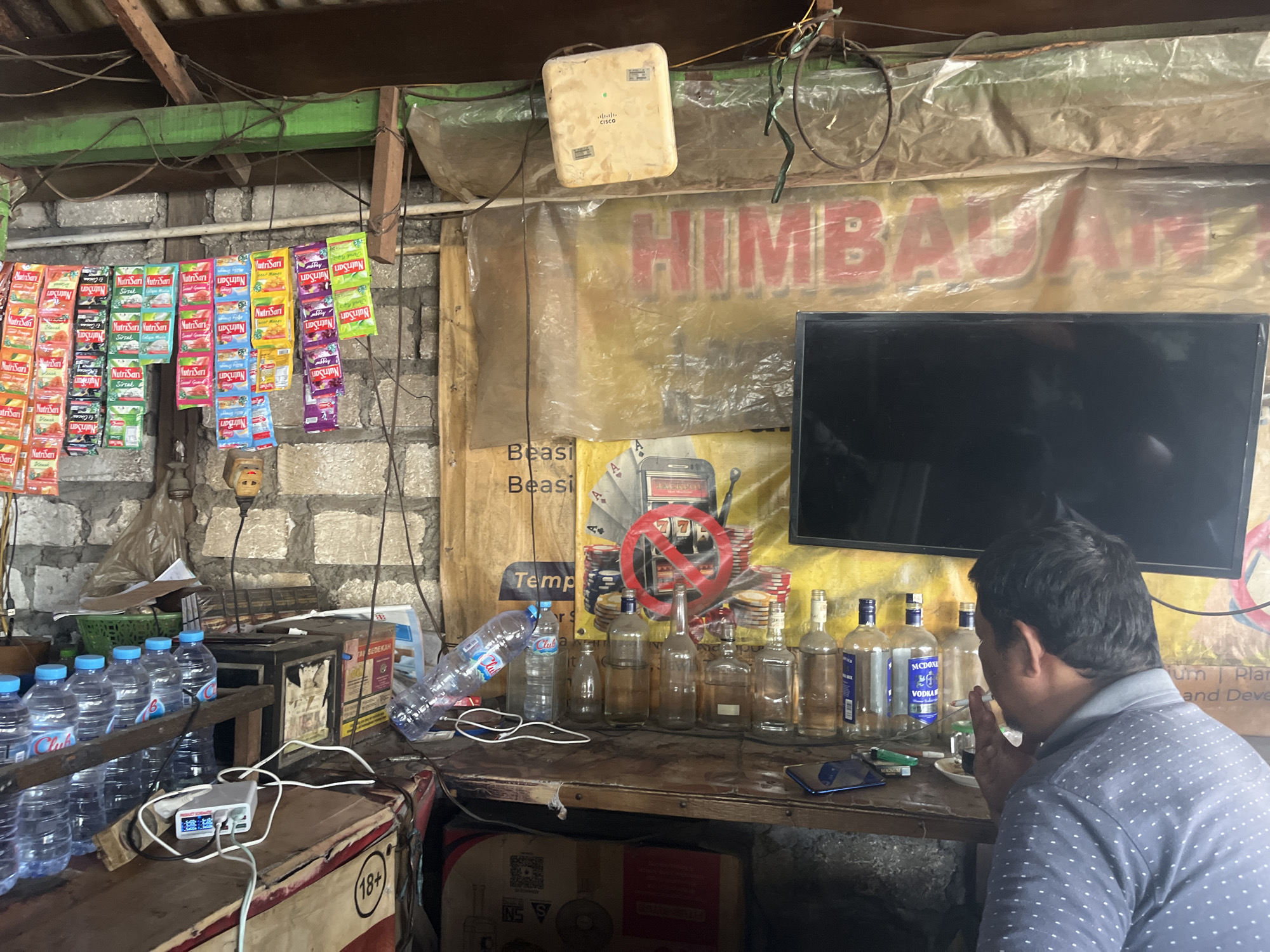 The image depicts an indoor stall, likely a small shop or warung, located in Dusun Metatu, Indonesia. The stall is cluttered with various items. In the foreground, a man with dark hair is seen from the back, wearing a blue and grey patterned shirt. He is smoking a cigarette and appears to be looking towards the right of the frame.
On the counter in front of him, there are several empty glass bottles, some with labels, including "McDONALD'S VODKA." There are also some smaller bottles and a smartphone lying on the counter. To the left, a row of bottled water is visible, along with a multi-port USB charger connected to wires.
The stall's background features a brick wall and a large yellow banner with red lettering, partially obscured by plastic sheeting. A black flat-screen television is mounted on the wall above the counter. To the left of the television, a beige Cisco router is attached to the ceiling. Hanging from the ceiling are various cables and wires.
Further to the left, numerous small colorful packets of what appear to be instant drink mixes or snacks are displayed, hanging in rows. These packets have labels such as "NutriSari". A green plastic basket is also visible in this area. The overall impression is a busy, utilitarian space where goods are sold.