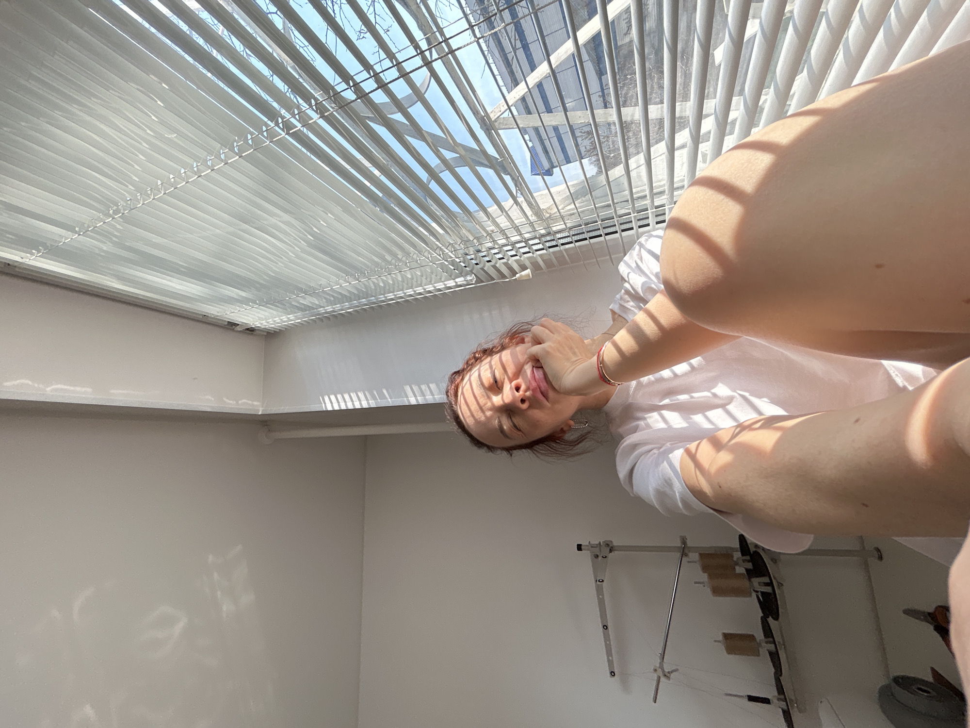 The image, taken from a low angle, shows a woman in a white shirt sitting near a window with closed blinds, filtering sunlight into the room. Her hand is touching her face. Behind her, a white wall and a clothes drying rack are visible. The scene is indoors. The location and city cannot be determined from the image.