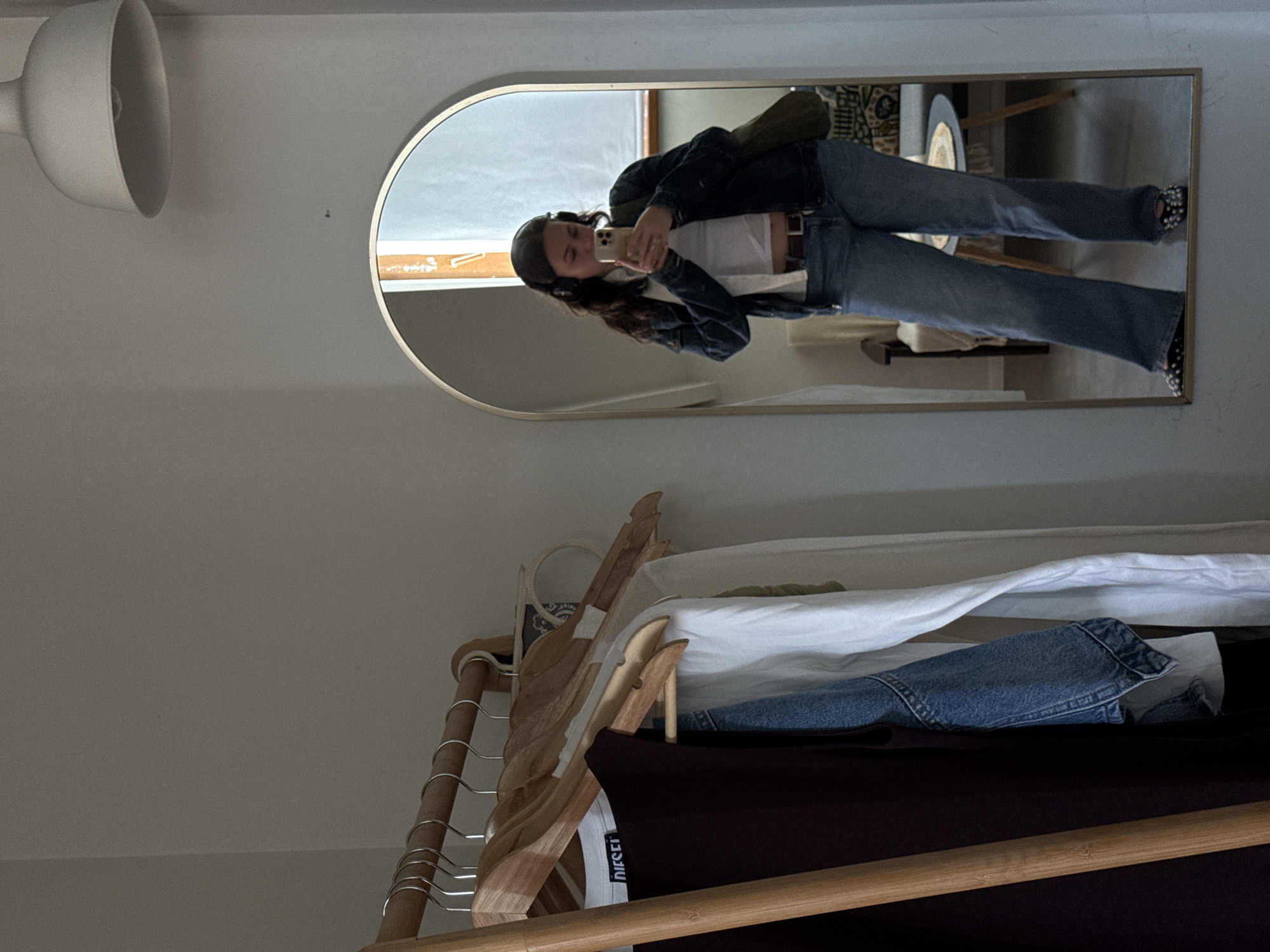 A woman is taking a mirror selfie in what appears to be an indoor space. The mirror is an arched rectangular shape with a gold frame. The woman is wearing a dark jacket, white top, and jeans with patterned shoes. She is holding a smartphone and looking at the screen. The mirror reflects a room with what seems to be a clothing rack in the foreground. The clothing rack holds hangers and various garments, including a dark shirt and a white shirt.
Based on the context provided, there's no explicit information within the image to determine the location in Yerevan, Armenia. The scene portrays a domestic interior without any readily identifiable external landmarks, symbols, or cityscapes that would suggest a specific location within Yerevan.