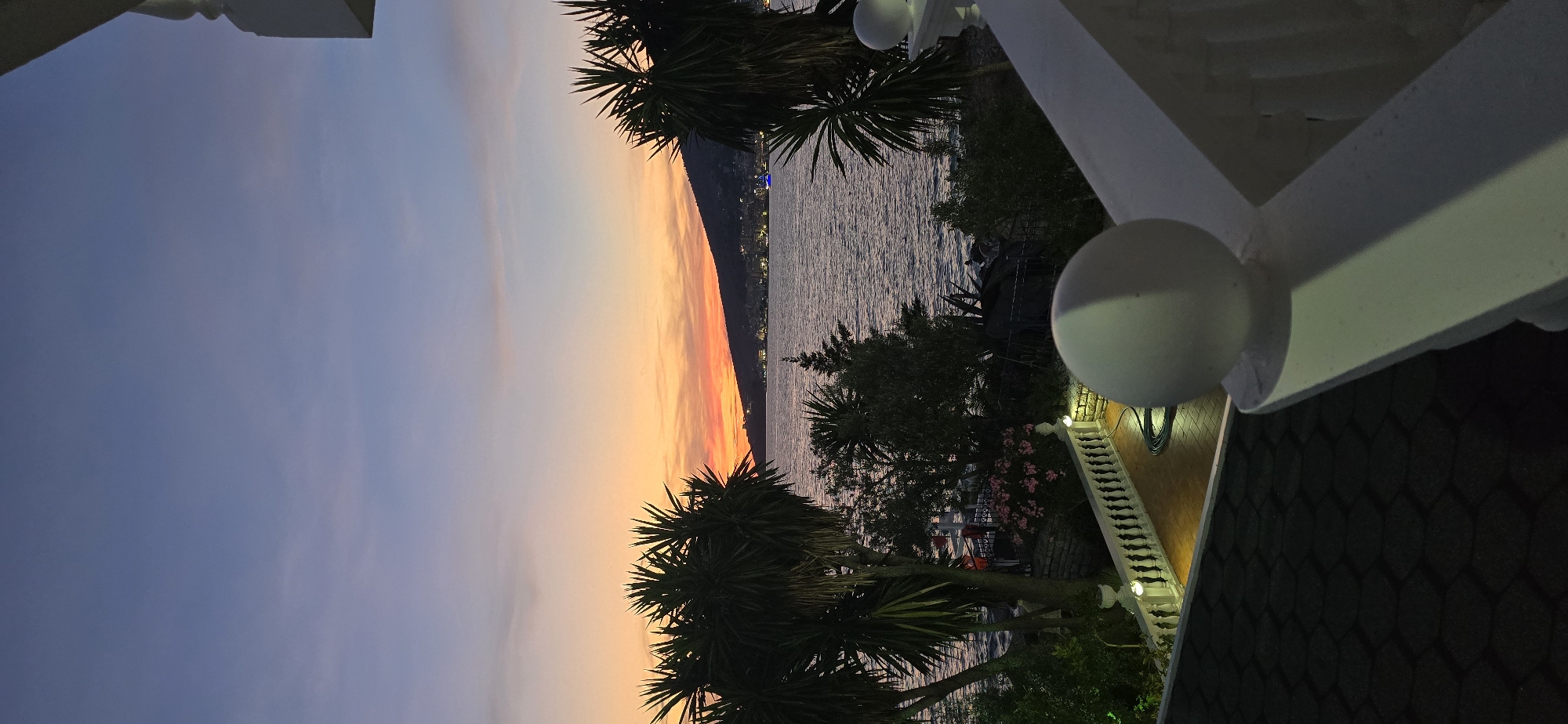 The image depicts a scenic view from a balcony overlooking the sea at sunset. The sky transitions from a deep blue at the top to vibrant orange and pink hues near the horizon, where a coastal town is visible with illuminated buildings and a blue boat in the water. Palm trees and lush vegetation frame the scene, suggesting a warm climate. A white railing with decorative balls and a patterned roof are in the foreground, indicating a residential or hotel property. The overall atmosphere is tranquil and picturesque. The location is identified as Saranda, Albania.