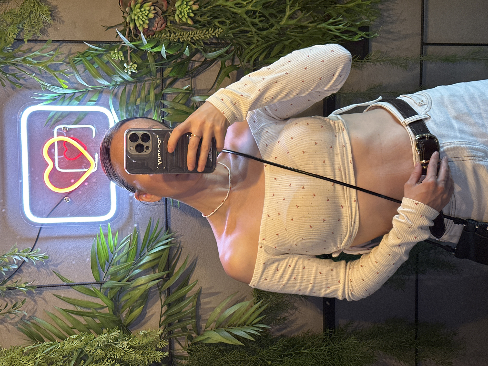 A person is taking a selfie in front of a decorated wall. The wall features various artificial green plants and a neon sign with a heart shape in red and yellow. The person is wearing a patterned long-sleeved crop top, white pants with a brown leather belt, and a delicate beaded necklace. They are holding their phone with their left hand, and the phone case is dark. The exact location within the city of Votkinsk, Russia is not discernible from the image.