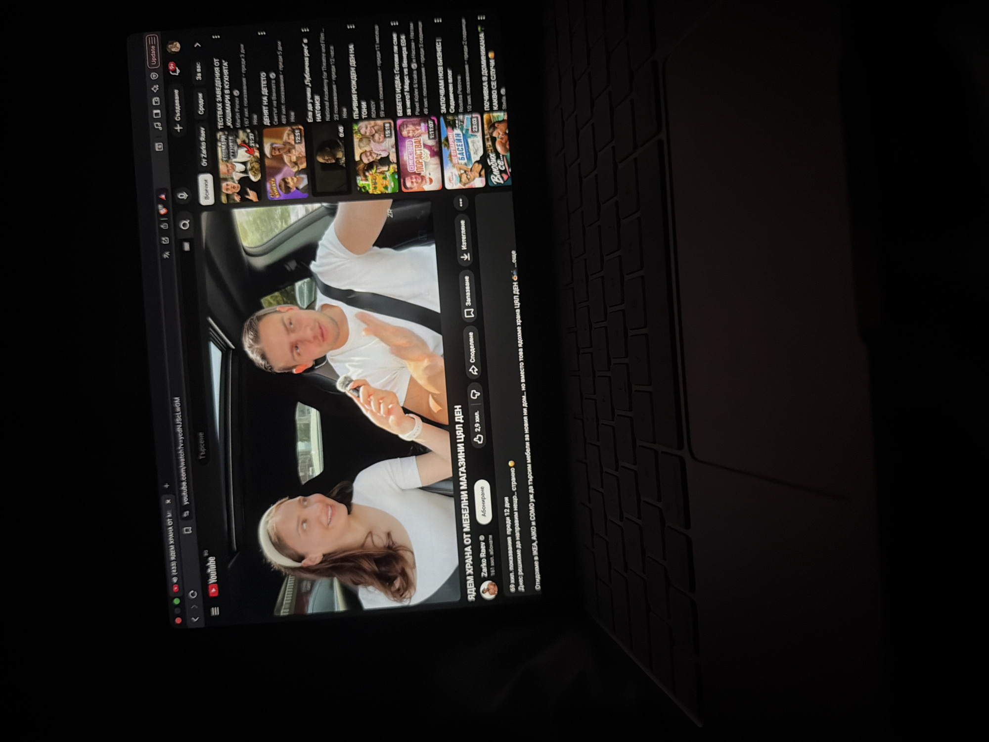 The image captures a laptop screen displaying the YouTube website in a dimly lit setting, likely at night or indoors. The main video playing shows a man and a woman, both dressed in white tops, inside a car. The man is in the foreground, holding an object, while the woman behind him smiles.
The video is titled "ЯДЕМ ХРАНА ОТ МЕБЕЛНИ МАГАЗИНИ ЦЯЛ ДЕН" (WE EAT FOOD FROM FURNITURE STORES ALL DAY) by channel "Zarko Raev," which has 161k subscribers. It garnered 69k views 12 days ago. The video's description snippet mentions, "Отидохме в ІКЕА, АЈКО и СОМО уж да търсим мебели за новия ни дом... но вместо това ядохме храна ЦЯЛ ДЕН..." (We went to IKEA, AIKO and SOMO supposedly to look for furniture for our new home... but instead we ate food ALL DAY...).
On the right side of the screen, several recommended videos are visible, including:
* "ТЕСТВАХ ЗАВЕДЕНИЯ ОТ „КОШМАРИ В КУХНЯТА" (I TESTED ESTABLISHMENTS FROM "NIGHTMARES IN THE KITCHEN") by Martin Penev (21:37).
* "ДЕНЯТ НА ДЕТЕТО" (CHILD'S DAY) by Светът на Ванката (The World of Vankata) (12:51).
* "Ела да учиш Лублична реч в НАТФИЗИ" (Come to study speech in NATFIZ) by National Academy for Theatre and Film (0:40).
* "ПЪРВИЯ РОЖДЕН ДЕН НА Тони" (TONY'S FIRST BIRTHDAY) by RONSY.
* "БЕБЕТО ИДВА: Готовили сме за него? Марс vs Венера Е04" (THE BABY IS COMING: Have we prepared for it? Mars vs Venus E04) by Pavel Kolev & Icakа и Насам Натам (15:18).
* "ЗАПОЧВАМ НОВ БИЗНЕС | Седмичен влог" (I'M STARTING A NEW BUSINESS | Weekly Vlog) by Rositsa Petrovs (23:03).
* "ПОЧИВКА В ДОМИНИКАНА КАКВО СЕ СЛУЧИ" (VACATION IN DOMINICAN REPUBLIC WHAT HAPPENED) by Stella (1:11:07).
The browser's address bar shows "youtube.com/watch?v=ycdNJScLWGM" and the word "Търсене" (Search). The laptop's keyboard is covered with a dark, textured protector. The image is taken at an angle, focusing on the screen content.