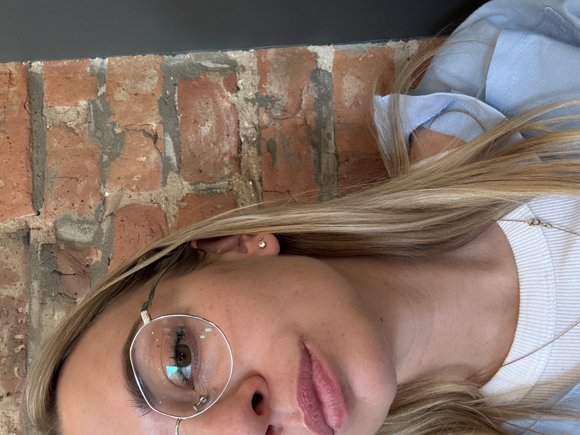 A partially visible woman with blond hair is angled downwards, wearing round glasses and light pink lipstick. She is wearing a white ribbed top and a light blue shirt. She has a small earring in her ear. To the left, a brick wall is visible. Above the brick wall is a dark grey surface. I cannot determine the location in Krasnoyarsk, Russia from this image.