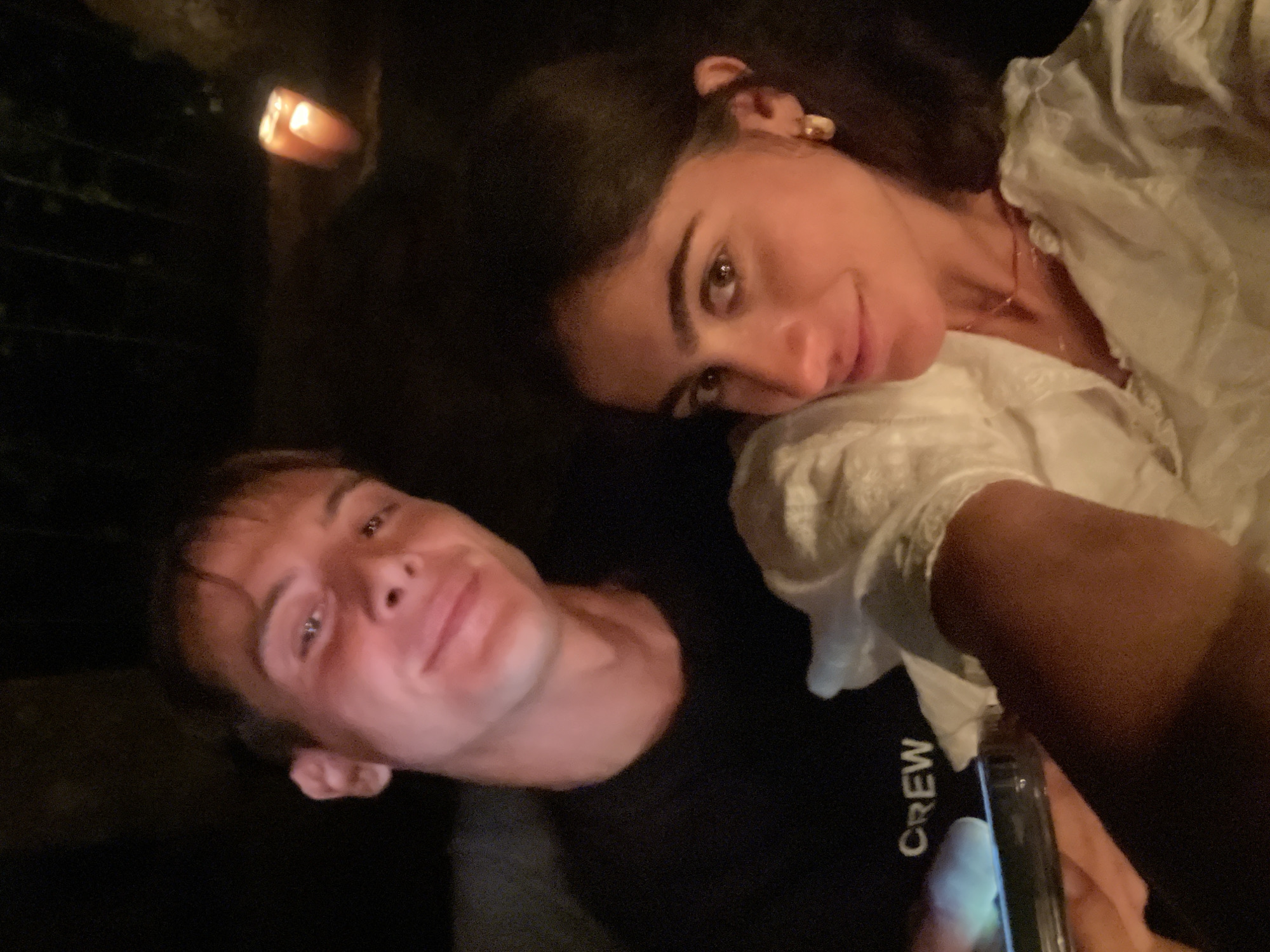 The image shows a close-up selfie of two young adults, a man and a woman, seated outdoors at night. The man is on the left, wearing a black t-shirt with "CREW" printed on it, and is holding a smartphone. The woman is on the right, wearing a white top with a ruffled neckline and a delicate necklace. Both are smiling. The background is dark and out of focus, but a lit candle can be seen in the upper right corner. The overall atmosphere is intimate and casual, suggesting they are on a date or spending time together. The setting appears to be a garden or patio area, with hints of greenery in the background. Vila Nova de Cerveira, Portugal, is known for its natural beauty and charming atmosphere, making it a plausible location for such a scene.