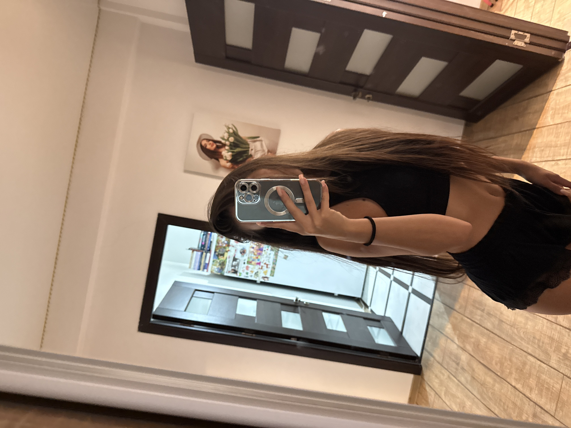 The image shows a young woman taking a selfie in a hallway with wooden floors and two dark brown doors. One door is partially open, revealing a room with a tiled floor and shelves filled with books and various items. On the wall opposite the open door, there is a framed picture of a woman wearing a hat and holding tulips. The woman in the selfie is wearing a black crop top and black shorts. Her long brown hair is down, and she is holding a smartphone to take the picture. The environment appears to be an indoor residential setting. There is no visible information to suggest the specific location within Chișinău, Moldova.