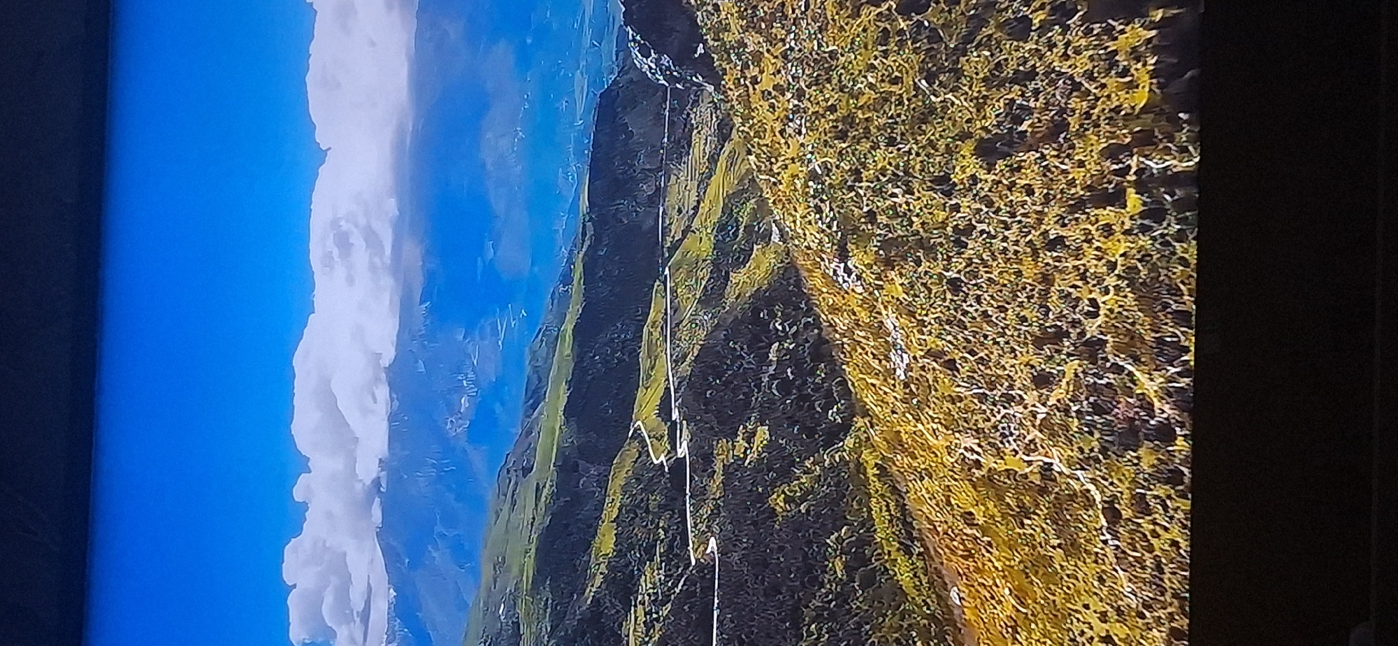 The image depicts a sweeping, mountainous landscape under a bright blue sky with scattered white clouds. A winding road snakes its way through the green and yellow-tinged hills in the foreground and midground. In the distance, a range of larger, blue-hued mountains is visible, partially obscured by clouds and haze. The terrain is rugged and appears to be characterized by steep slopes and valleys. The overall scene conveys a sense of natural grandeur and remoteness. There is no indication of the city of Bila Tserkva, Ukraine, in this image.
