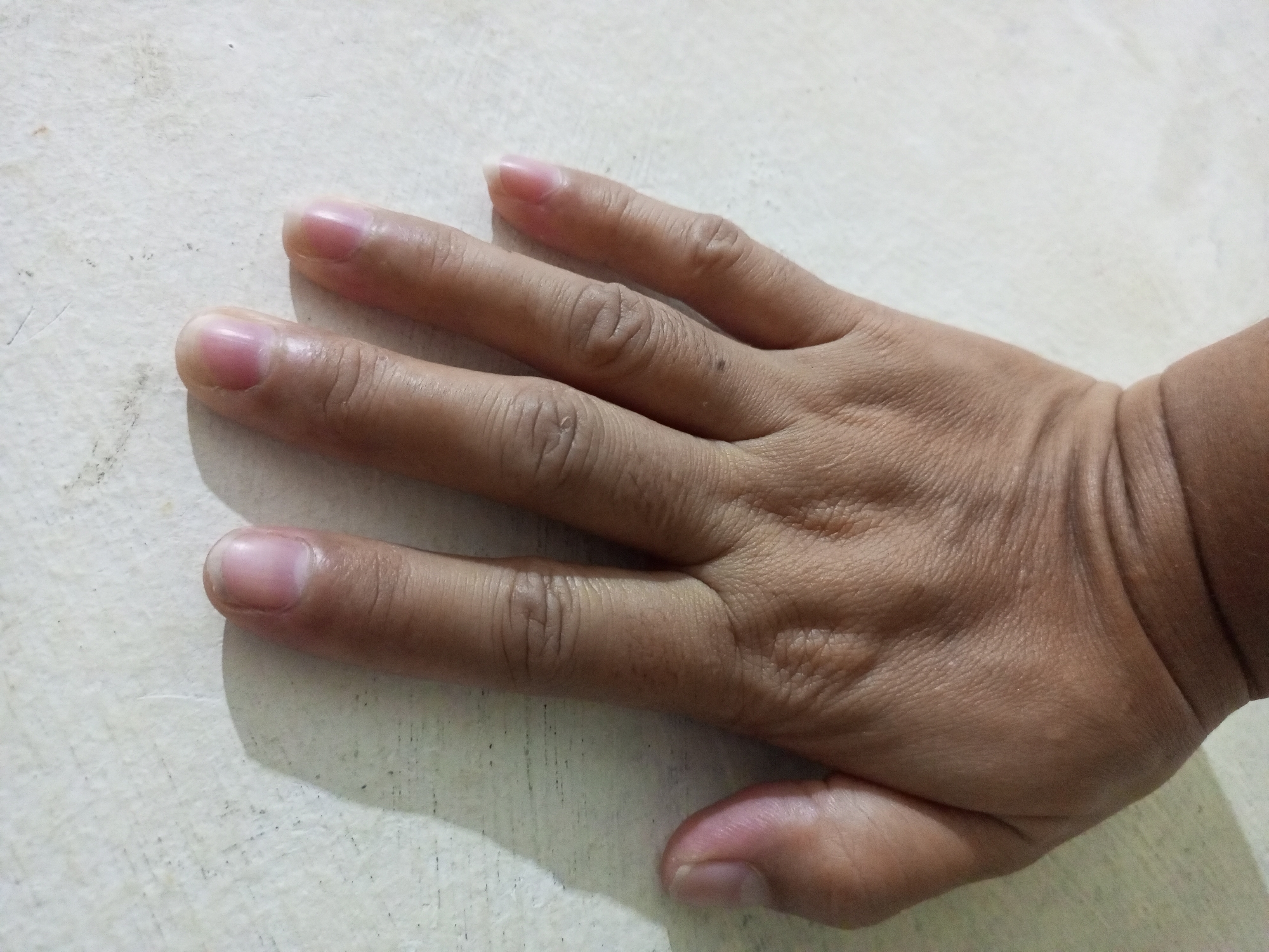 A close-up, overhead shot shows a person's left hand, palm down, resting on a light-colored, textured surface. The hand is positioned with the fingers spread and slightly curled, extending from the bottom right to the top left of the frame. The skin appears to be of a medium tone, with visible wrinkles on the knuckles and the back of the hand. The fingernails on the four extended fingers are short and rounded, with a healthy pinkish hue. The thumb is bent at the base of the hand, its tip visible near the bottom right corner. A faint shadow is cast by the hand onto the surface to the left. The surface itself has a subtle grainy texture and appears to be made of a solid material, possibly wood or laminate. No other objects or individuals are present in the image.