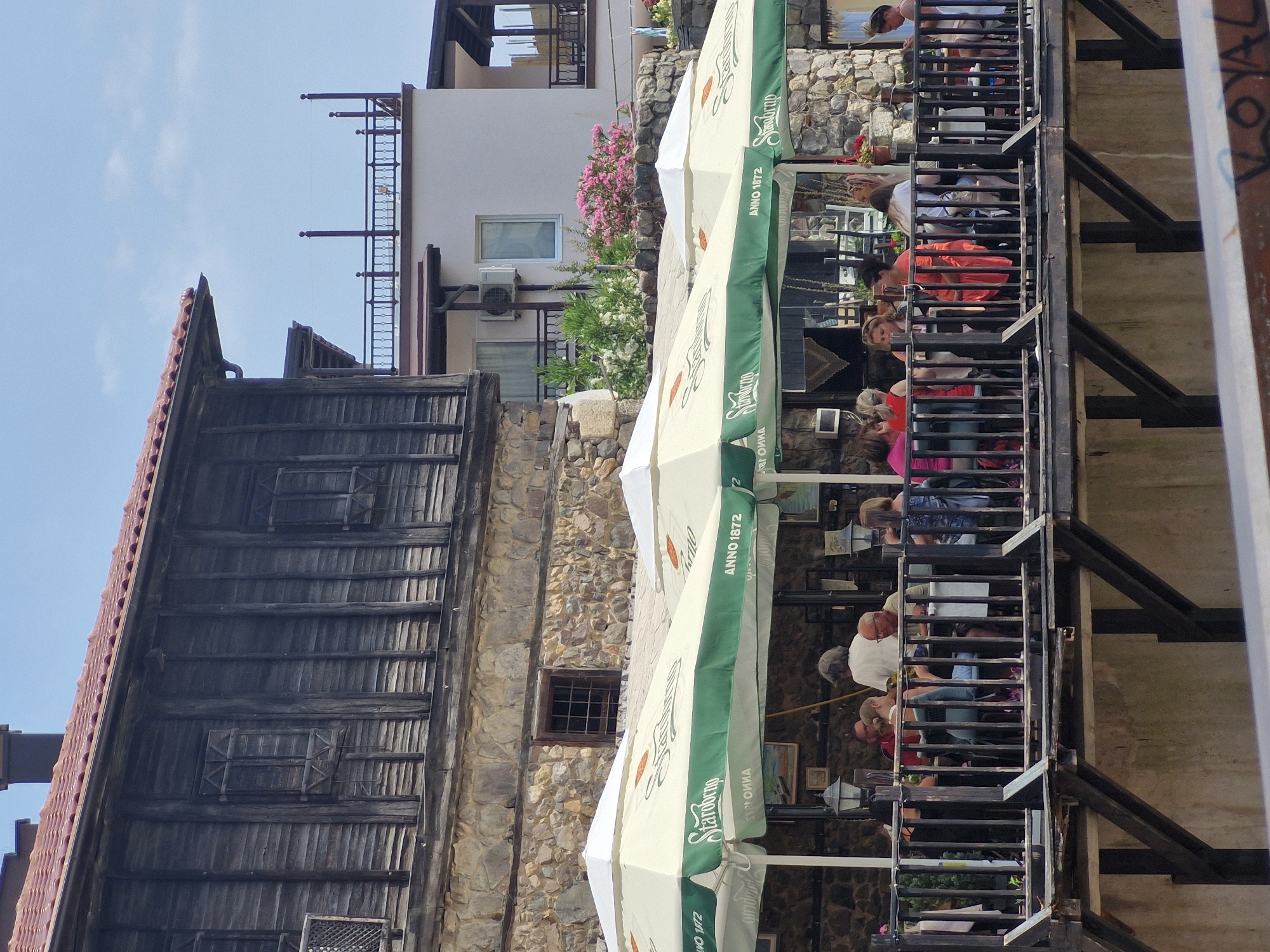 The image captures an outdoor seating area of a restaurant in Sozopol, Bulgaria, situated along a waterfront or canal. The restaurant is adorned with large, cream-colored umbrellas featuring green trim and the "Staropramen" logo, along with the text "ANNO 1872". Several people are seated at tables, engaged in dining or conversation. The architecture visible includes a traditional wooden building with stone foundations and a white building with balconies and an air conditioning unit. Lush pink and white flowering bushes are planted in front of the white building. The foreground shows a wooden railing and supports of the elevated restaurant deck. The sky is a clear blue with a few white clouds.