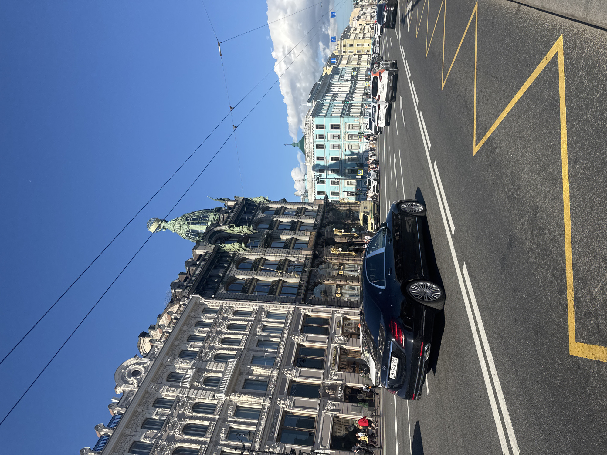 This media file captures a street scene in Saint Petersburg, Russia, on a bright, sunny day with a clear blue sky and scattered white clouds. The perspective is from street level, looking up towards the ornate architecture of historic buildings lining a wide road.
The primary subjects are the buildings themselves, showcasing intricate neoclassical facades with numerous windows, balconies, and detailed sculptural elements. One prominent building is a light blue structure with a green dome. To the left, a darker, more elaborately decorated building features a large dome topped with a globe-like structure.
The scene depicts a busy urban environment. Several cars are visible on the road, including a black Mercedes-Benz sedan in the foreground and other vehicles further down the street, suggesting moderate traffic. Some people can be seen walking on the sidewalks and near the buildings, indicating that the city is active. The road has clear lane markings, including double white lines and yellow zig-zag lines, likely indicating a no-parking zone or a pedestrian crossing area.
The time of day appears to be daytime, given the bright sunlight and distinct shadows cast by the buildings. The weather is pleasant and fair. Electrical wires are strung across the sky, a common sight in urban settings.
There is visible text on the license plate of the black Mercedes-Benz sedan, which reads "E875PX 198". Some shop signs are also faintly visible on the buildings, though they are not clearly legible. The overall impression is of a vibrant, historic European city captured on a beautiful day.