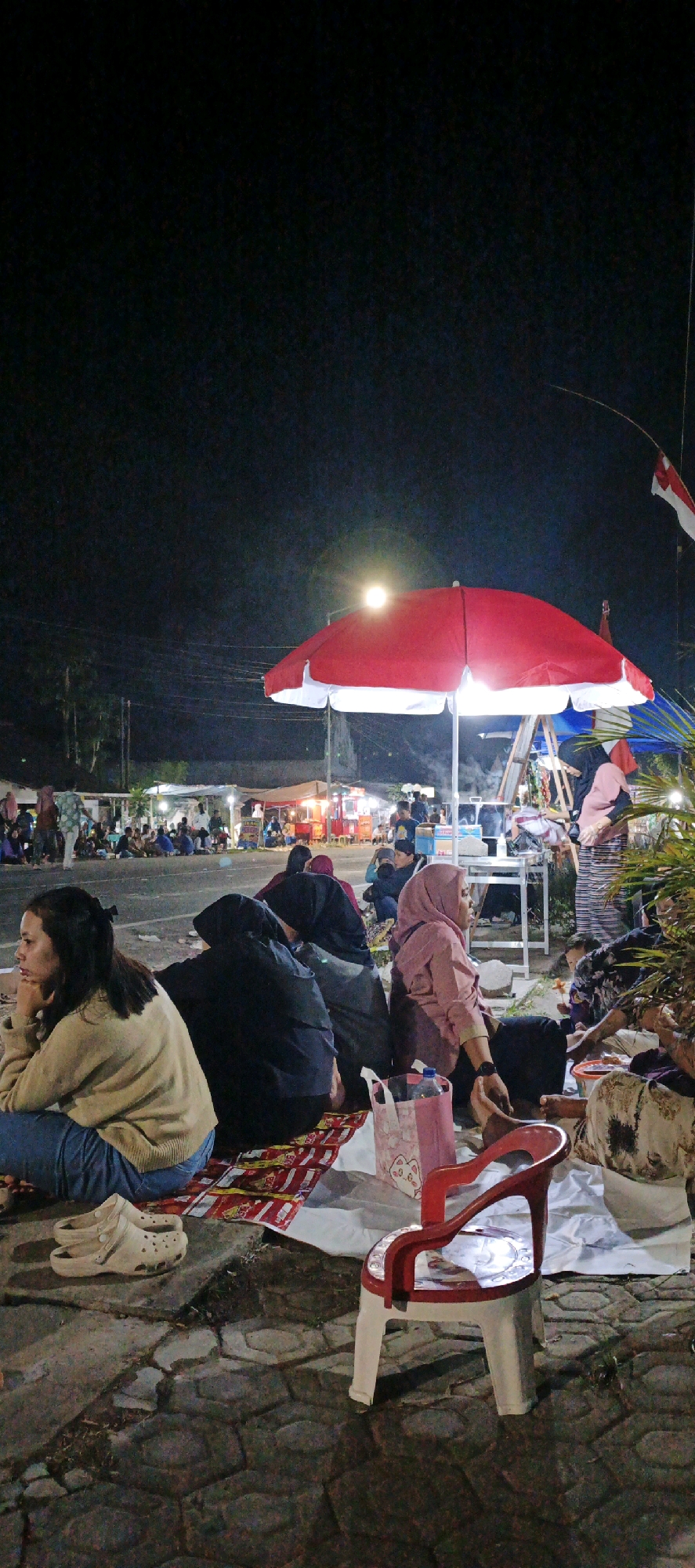 In Kasiyan, Indonesia, at night, a group of people are gathered outdoors. The scene appears to be a street or roadside market, with several food stalls or vendor booths visible in the background, illuminated by artificial lights. A large red and white umbrella is prominently featured in the foreground, providing shade.
In the immediate foreground, several individuals are seated on the ground, resting on mats or cloths. One person, a woman with dark hair tied back, is seated facing left, with her hand resting on her chin, appearing contemplative. She is wearing a light brown sweater and blue jeans. To her right, other individuals are seated, some wearing headscarves and traditional Indonesian attire. A red and white plastic chair is placed in the foreground, unoccupied. A pink and white patterned bag is visible near the seated individuals.
In the middle ground, more people are gathered around the vendor stalls, interacting and observing. The stalls appear to be selling food or other goods. A ladder leans against a structure behind the umbrella. Electrical wires are visible strung across the background. A flag, likely the Indonesian flag, is partially visible in the upper right corner of the image.
The ground is paved with interlocking hexagonal stones, suggesting a sidewalk or pedestrian area. The overall atmosphere is that of a lively night market or community gathering.