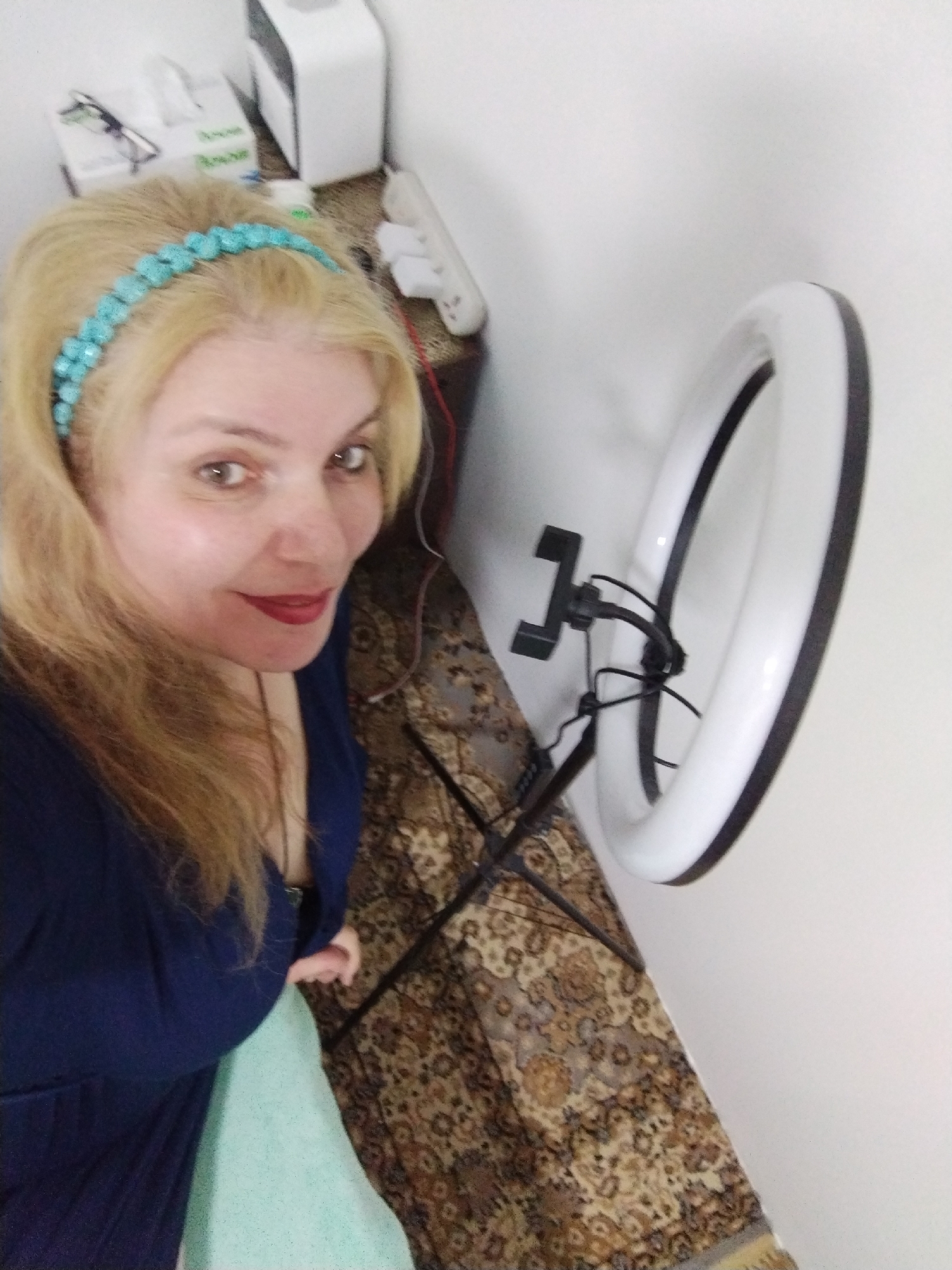 A woman with long blonde hair and a turquoise beaded headband is positioned on the left side of the frame, wearing a dark blue top and holding a pale green cloth. She appears to be in a room, possibly a bedroom, with a patterned rug on the floor. Beside her is a ring light mounted on a stand. In the background, there is a side table with a tissue box, glasses, and other items. The photo was likely taken in an interior setting, but the specific location in Al Mas'oudi, United Arab Emirates cannot be determined without additional information.