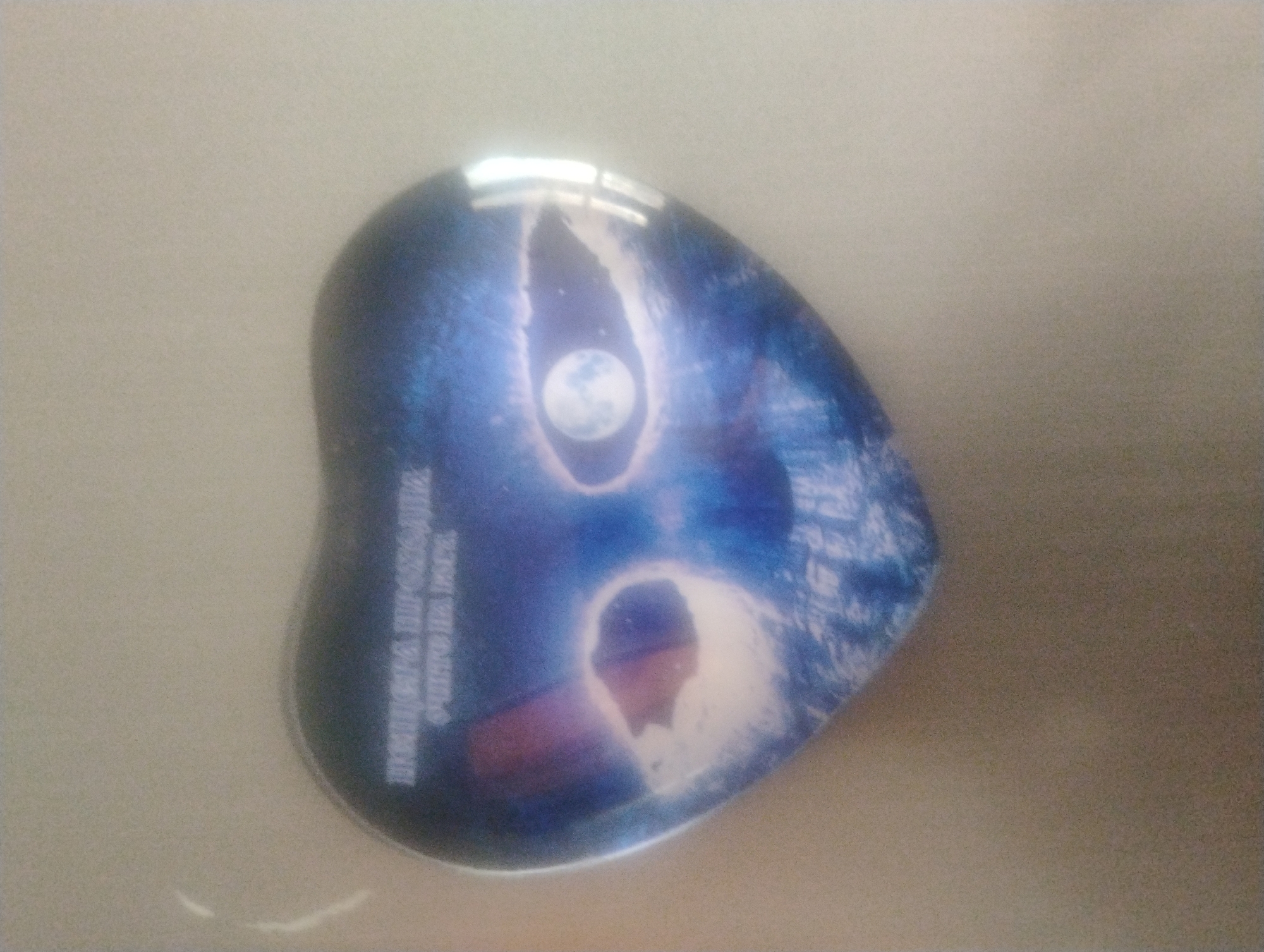 The image shows a heart-shaped magnet with a blue, outer space-themed design featuring two large, oval shapes that resemble eyes. Inside each eye-like shape, a celestial body is visible. The left eye contains what appears to be a reddish-purple nebula or planet fragment, while the right eye contains a full moon in a starry sky. Text in white Cyrillic script is superimposed at the top of the heart shape, reading "ПУШКИНА ПРОХОДНАЯ / ОЧИТОК НА ВОСК." The magnet is placed on a neutral-colored, possibly a wall or tabletop, with some subtle lighting reflections on its glossy surface. The image's quality is moderate, with some blurriness and graininess. The location specified, Rumyantsevo, Bulgaria, is not visually discernible from the provided image.