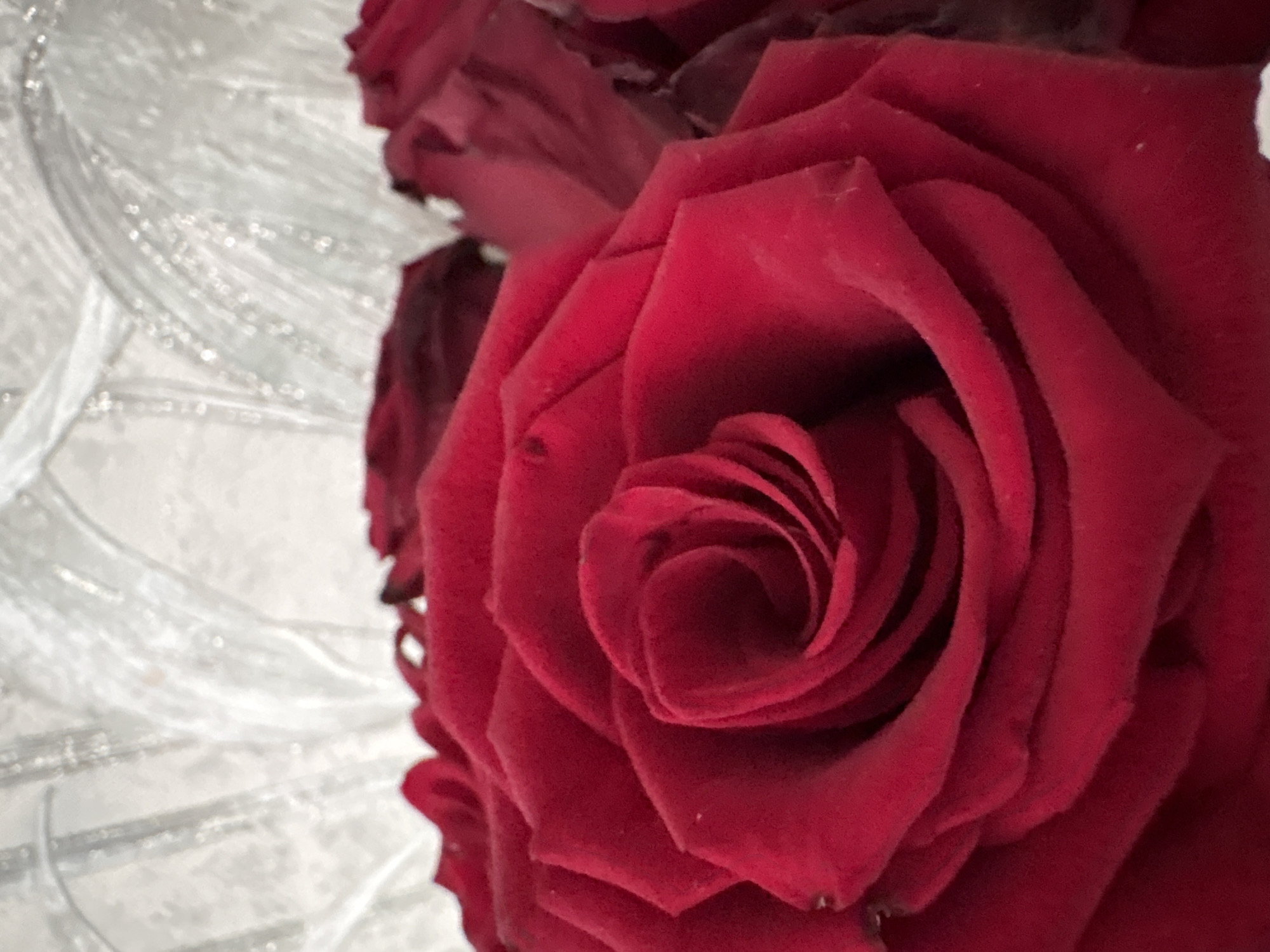 This close-up photograph features a bouquet of deep red roses, with one prominent bloom occupying the right side of the frame. The rose exhibits velvety petals, spiraling inwards towards its dark center. The rich, dark red hue of the petals is the dominant color in the image.
On the left side of the composition, a blurred background with a translucent or off-white appearance is visible. This background seems to be a textured decorative element, possibly fabric, a vase, or another surface, characterized by shiny, glittery, or reflective linear patterns and speckles.
The image is a still-life, focusing on the aesthetic details of the flowers. There is no discernible activity or event taking place. The lighting is soft, highlighting the textures of the rose petals and the reflective elements of the background. No specific time of day or weather conditions can be inferred, and there are no direct location cues or visible text within the image. The arrangement suggests an indoor setting.