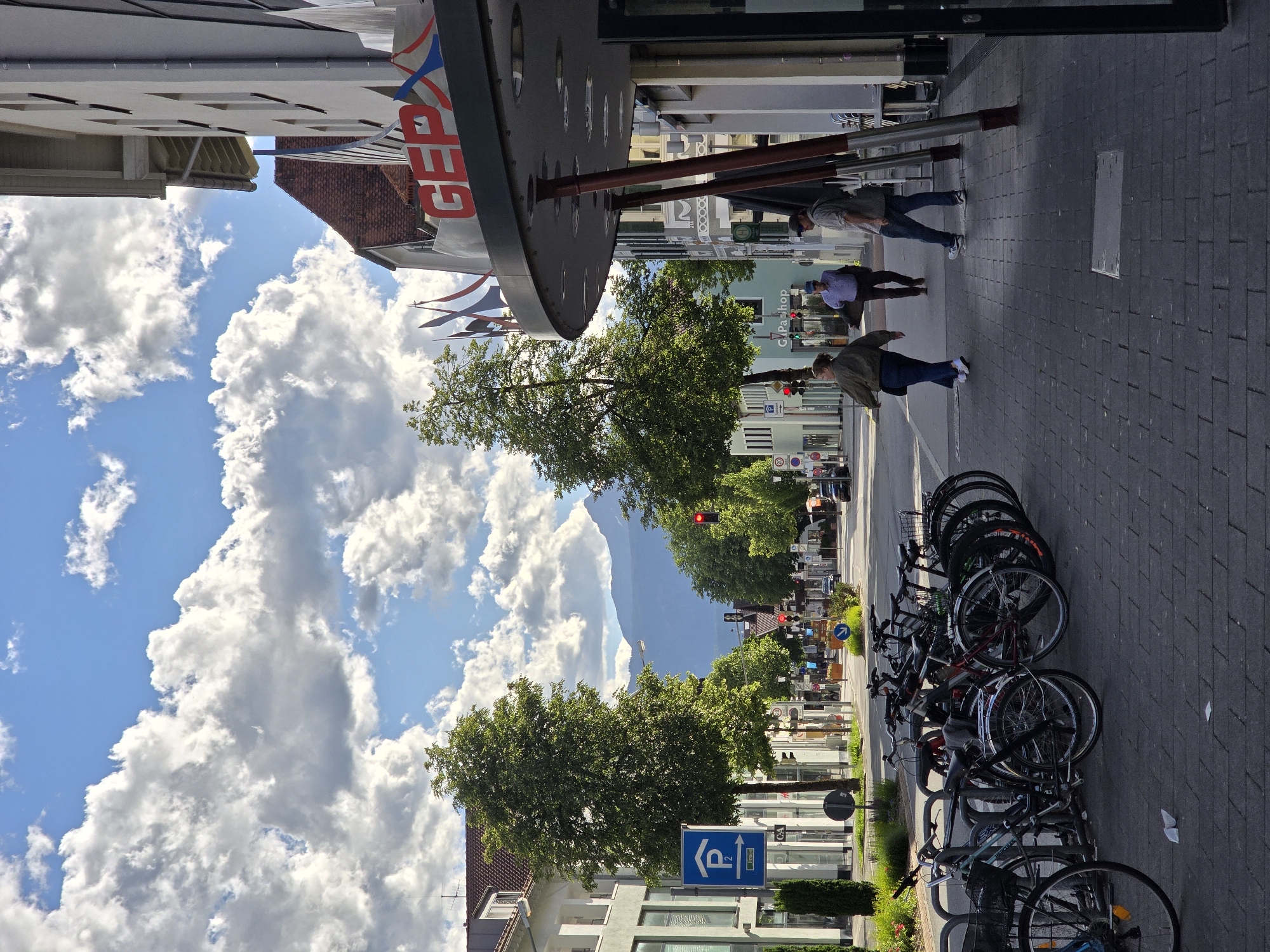 A street scene in Garmisch-Partenkirchen, Germany, features a paved sidewalk with several parked bicycles. A row of trees lines the street, and buildings are visible on either side. In the background, mountains rise against a sky filled with white, fluffy clouds. Three people are walking on the sidewalk. One woman in a green jacket and jeans is walking away from the camera. A man in a blue shirt and dark pants is walking toward the camera, and another man in a gray shirt and jeans is also walking toward the camera. A red traffic light is visible in the distance. The GEPX logo is displayed on the side of one of the buildings.