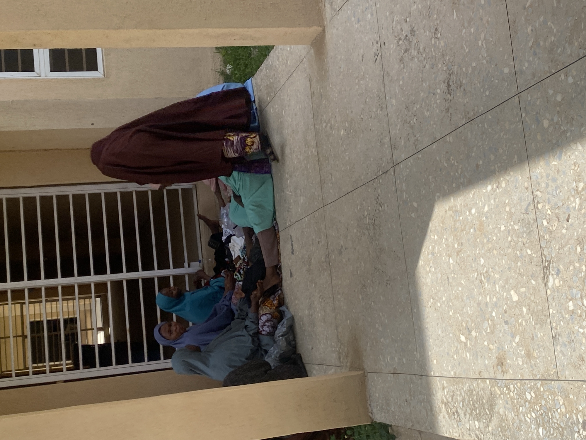 The image depicts a group of individuals, likely women, seated on the ground in an outdoor area. The setting appears to be a sheltered walkway or patio adjacent to a building with a barred window and a section of wall with a white-framed window. The ground is paved with large, speckled tiles.
Several individuals are visible from the waist up, all wearing headscarves or hijabs. One person in the foreground is wearing a teal headscarf and appears to be looking to the right. Next to them, a person in a darker blue headscarf is also looking to the right. To the left of these two, a person in a light blue headscarf is looking forward. The person furthest to the left, also wearing a light blue headscarf, is facing away from the camera. In the background, a person is seated, largely obscured by a dark maroon fabric draped over their head and shoulders. This person is wearing a teal garment underneath.
Various items of clothing and bags are scattered on the ground around the seated individuals. The environment suggests a casual gathering or waiting area. The lighting indicates it is daytime, with shadows cast across the tiled floor. The overall scene is static, with no overt actions taking place beyond sitting and conversing or waiting.
The location is identified as Yali, Nigeria, suggesting an urban or semi-urban environment consistent with the architectural features observed.