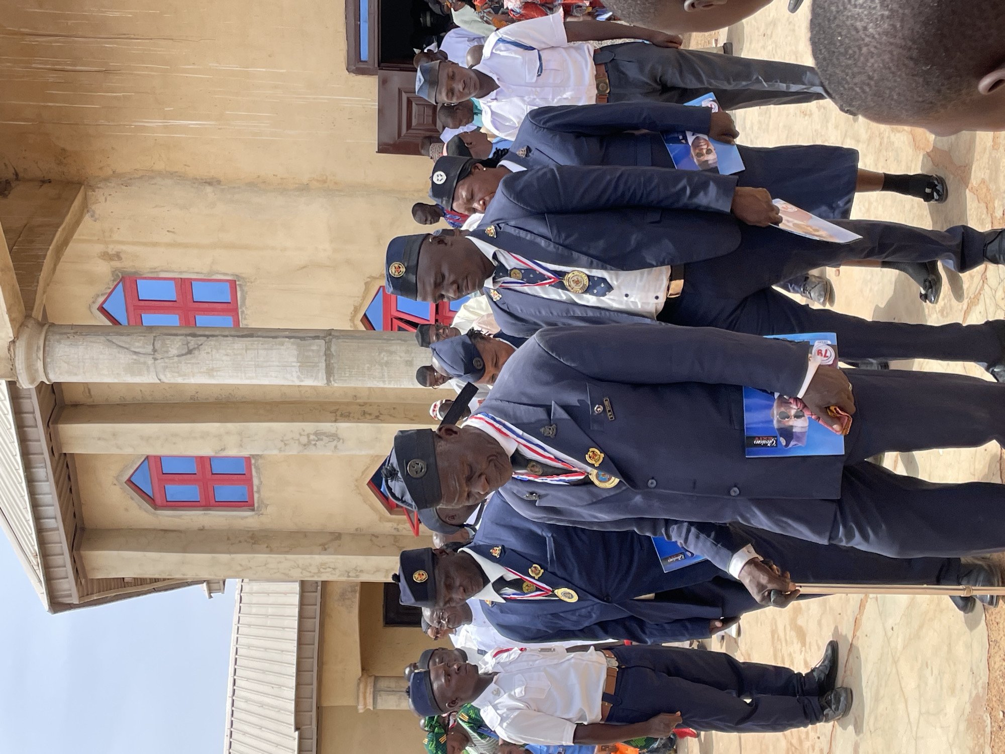 The image depicts a formal outdoor gathering of men in Ladura Foron, Nigeria, standing in front of a light brown building under bright daylight. Most of the men, predominantly older, are dressed in dark blue uniforms featuring suits, white shirts, ties, and hats, some adorned with medals and sashes in red, white, and blue. Other men wear white short-sleeved shirts, dark trousers, and caps. Their expressions are largely serious or contemplative.
Many attendees are holding small pamphlets or programs. Text visible on these includes "Global" and "Gideon" on one, which also features a photo of a man in sunglasses and a mask. Another program shows "TL" or "7L." At least two older men are using walking sticks. The building in the background has distinctive red-framed windows with blue panes, one with an arrow-like design, and features columns and cornices. The ground is a light-colored, possibly paved surface, with some dry earth visible. The clear, sunny weather suggests the event is taking place during the day. In the background, other individuals, some in traditional attire, are present, and a child's head is visible on the far right. The overall scene suggests an official ceremony, parade, or significant communal event.