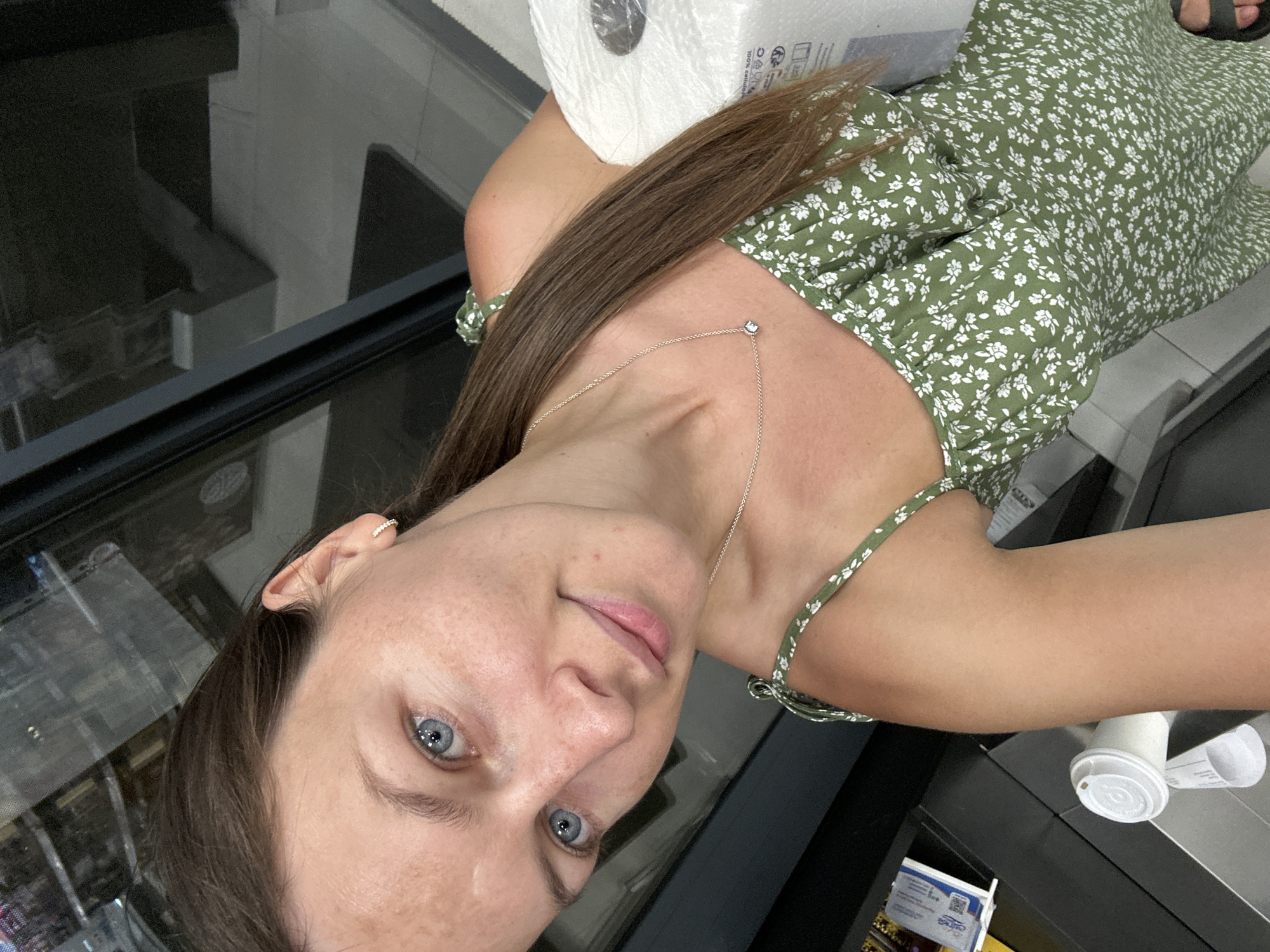 A woman with blue eyes and brown hair is taking a selfie. She is wearing a green floral dress and a necklace. She is holding a roll of paper towels. The background appears to be indoors, possibly a store or public restroom, with shelves and a window visible. The image is taken at an upward angle, showing the woman's face and upper body. The location of Donji Pobori, Montenegro is not visible in this image.