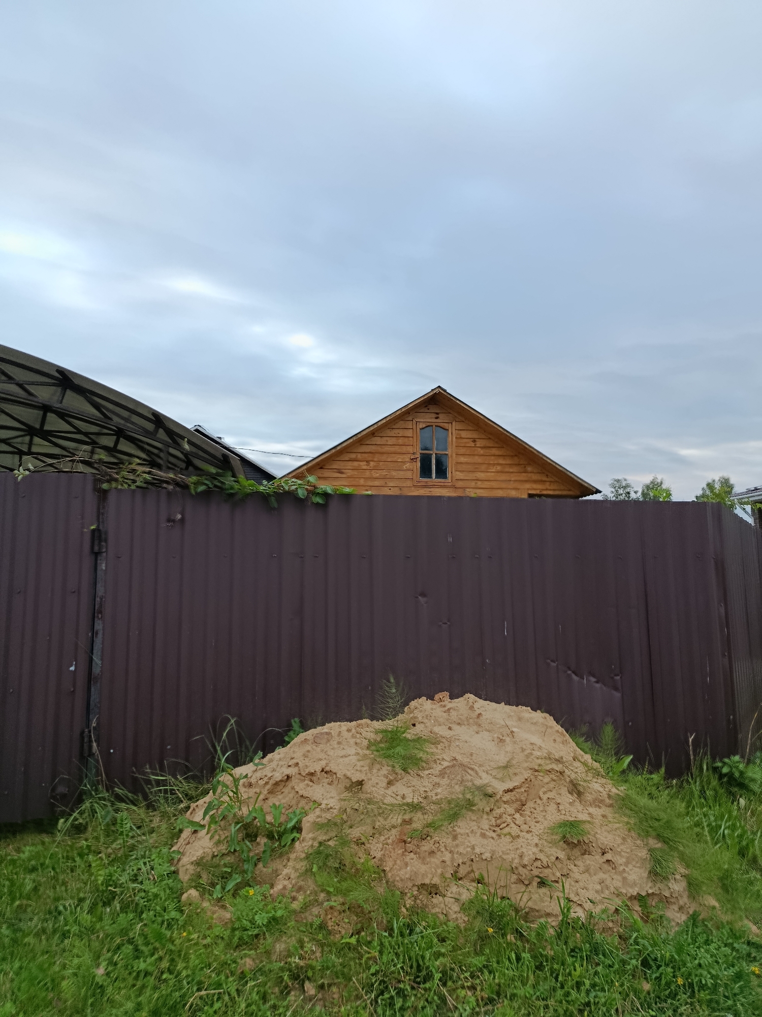 The image depicts a residential scene in Verеya, Russia. In the foreground, a mound of light-colored soil is visible amidst green grass and some sparse vegetation. Behind the soil mound, a tall, corrugated metal fence with a dark brown color extends across the frame. To the left of the fence, a section of a dark, translucent roof structure is present. Above the fence, the peak of a wooden house with a gabled roof and a small arched window is visible. The sky is overcast with muted tones of gray and white. The overall impression is one of a partially developed or rural property under a cloudy sky.